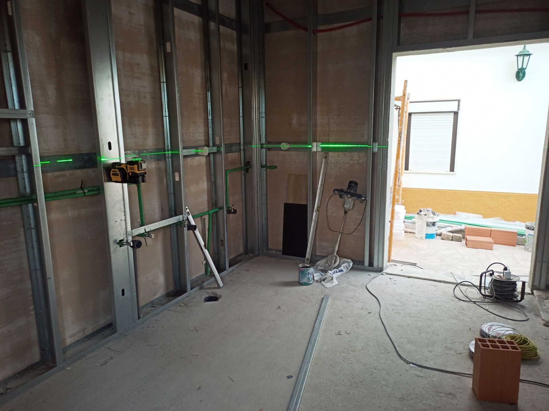 Interior em construção: Estrutura metálica com níveis a laser projetando linhas verdes. A porta dá acesso ao exterior.