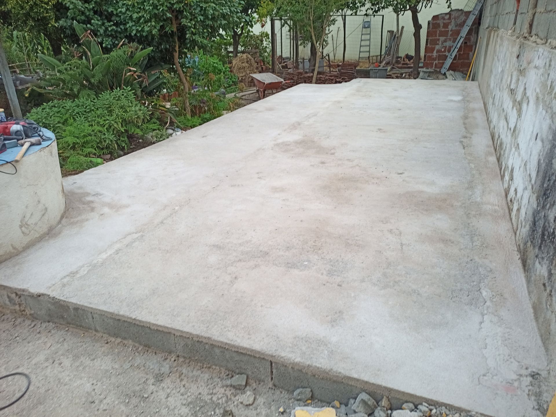 Placa de concreto em espaço externo, delimitada por um meio-fio baixo de concreto e um muro, cercada por folhagem.