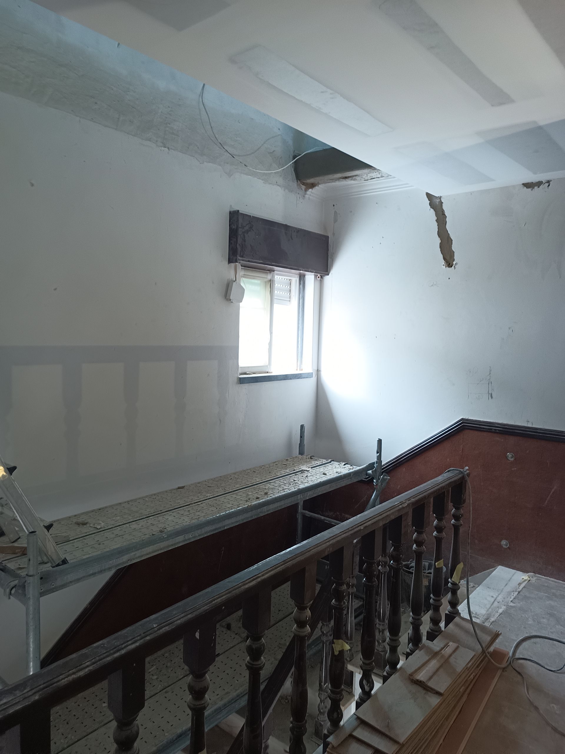 Foto do interior de uma escadaria em reforma; paredes brancas, corrimão de madeira, janela, andaimes e teto danificado.