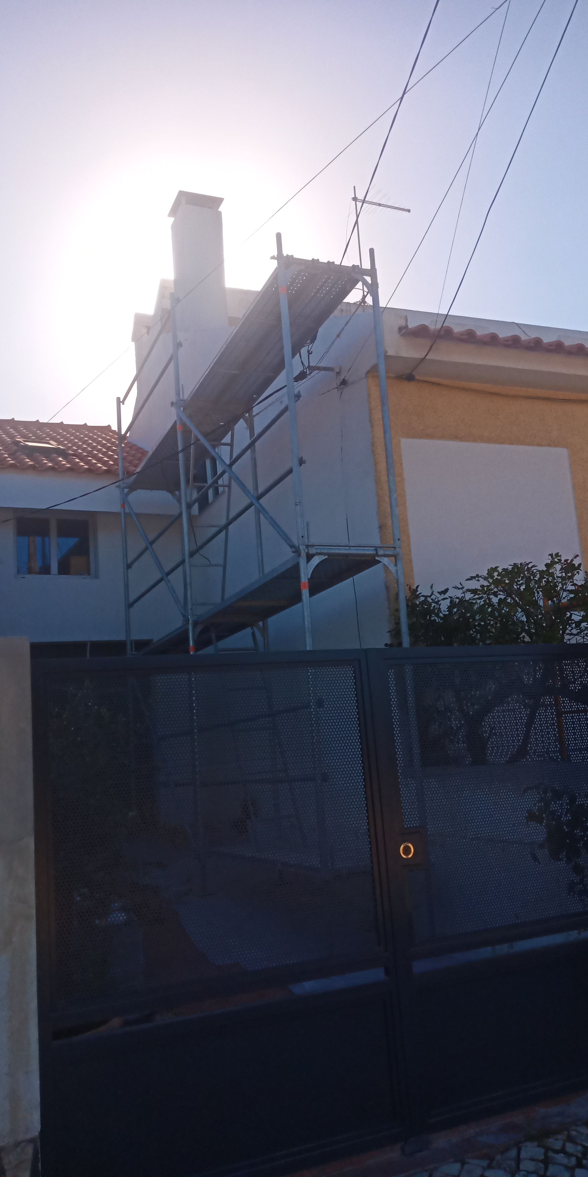 Andaimes em um prédio com chaminé; um portão preto em primeiro plano. Sol forte.