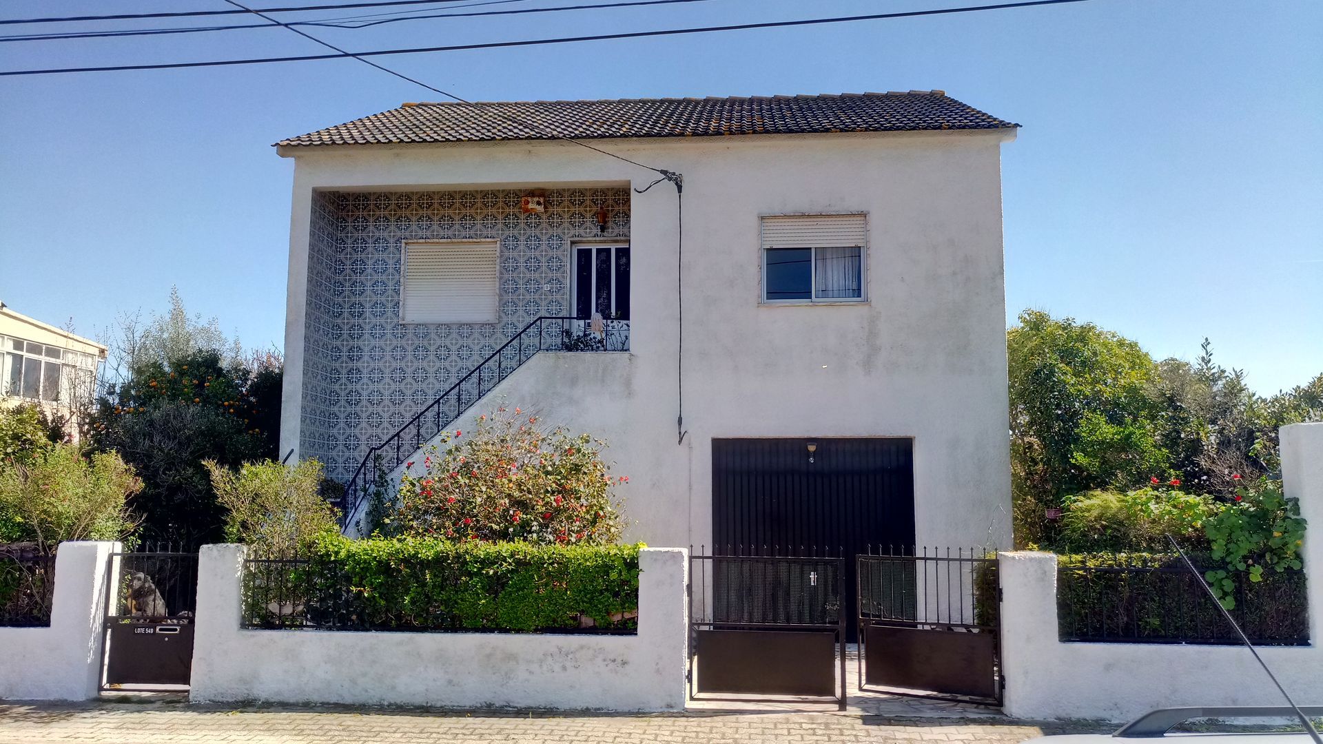Casa branca de dois andares com portão preto e telhado de telhas, árvores de ambos os lados contra um céu azul.