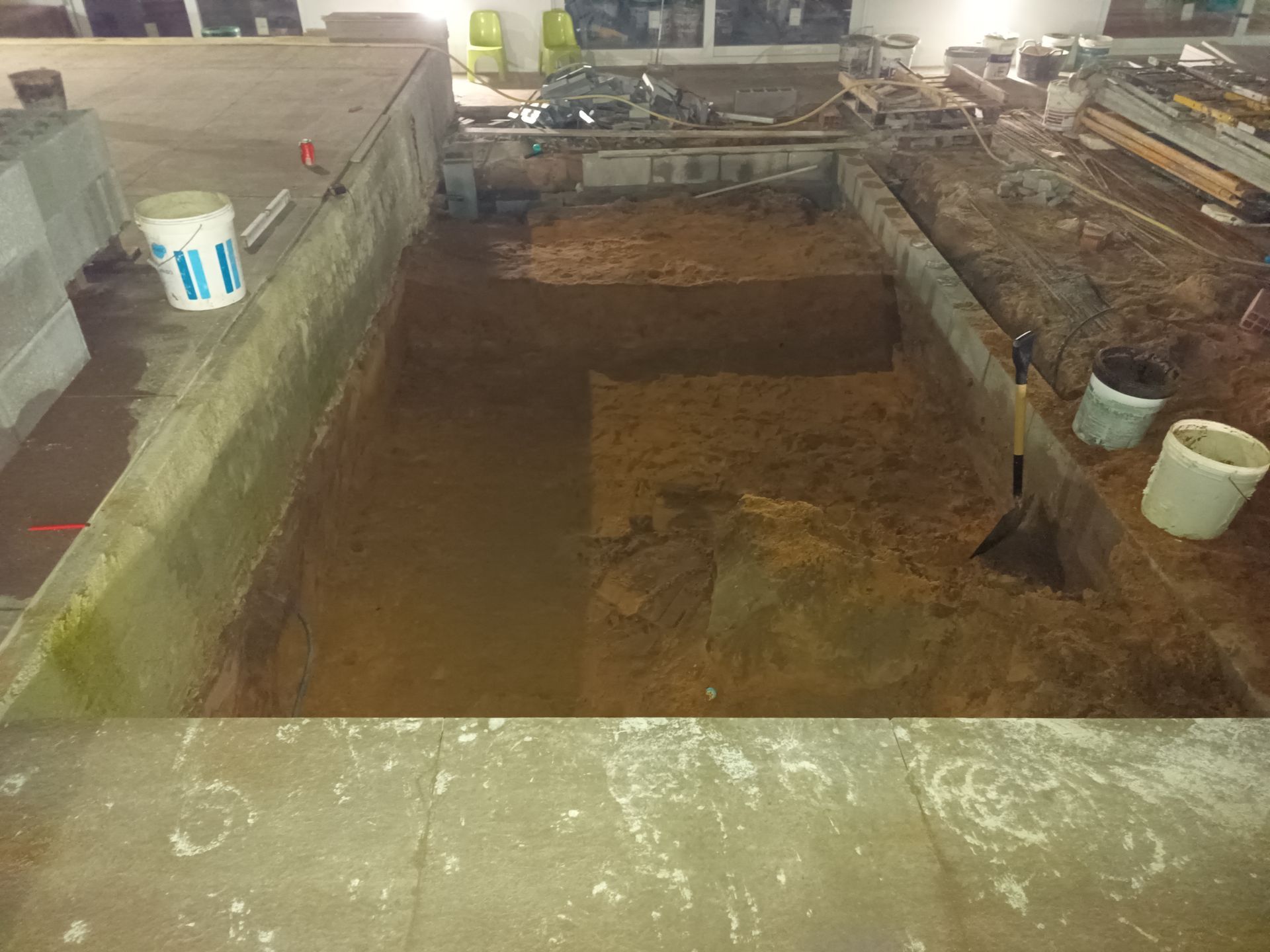 Canteiro de obras com grande escavação em piso de concreto. Há baldes e ferramentas no local.