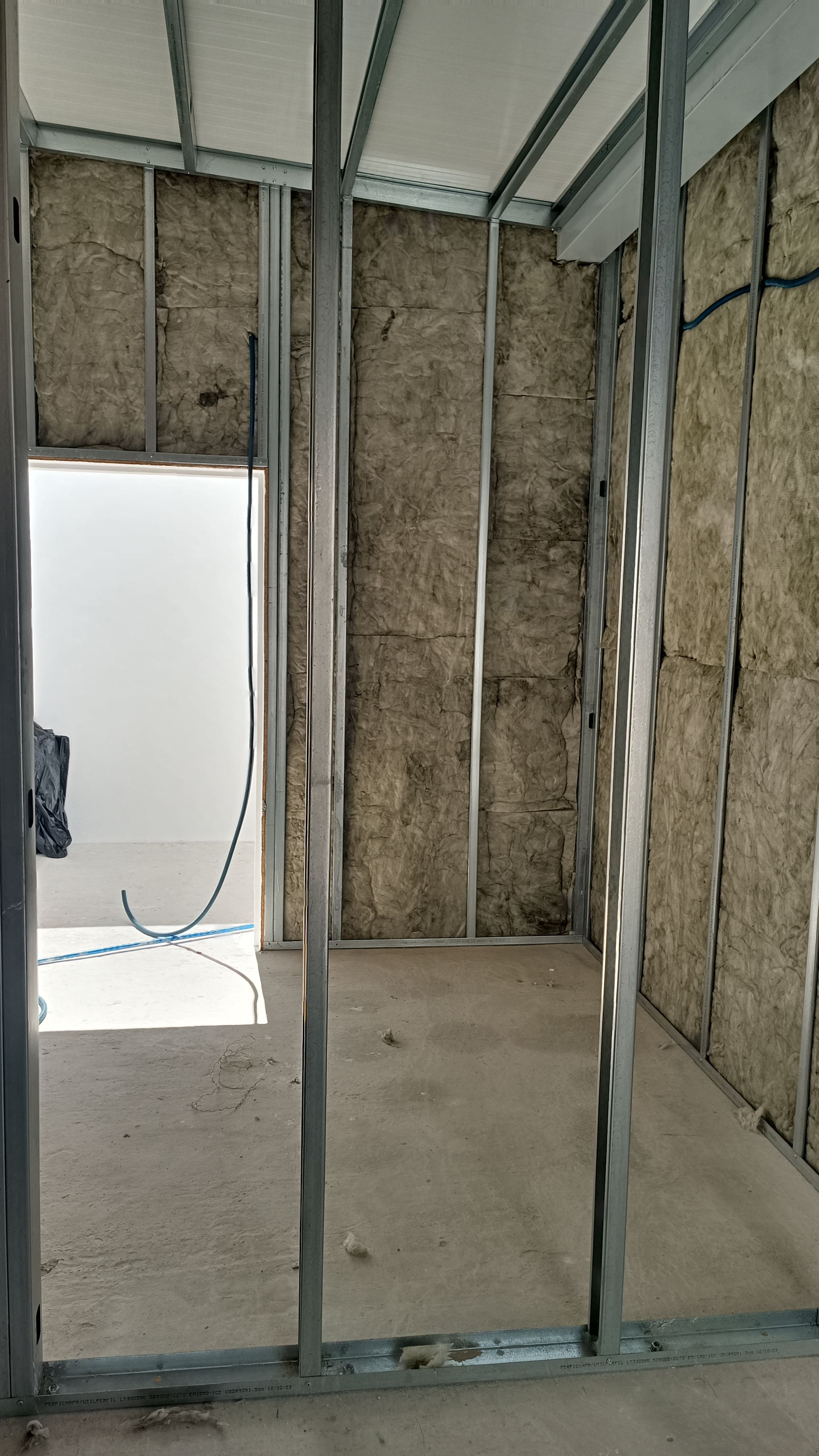 Vista interna de um cômodo em construção, mostrando a estrutura metálica, o isolamento e a porta.
