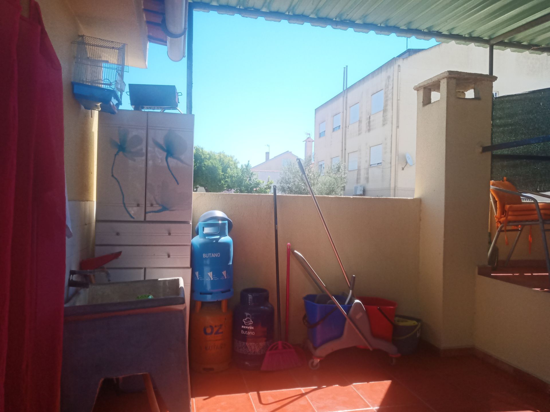 Área externa coberta com vista para os prédios. Inclui botijões de gás propano, espaço para armazenamento e materiais de limpeza. Ensolarada e bem iluminada.