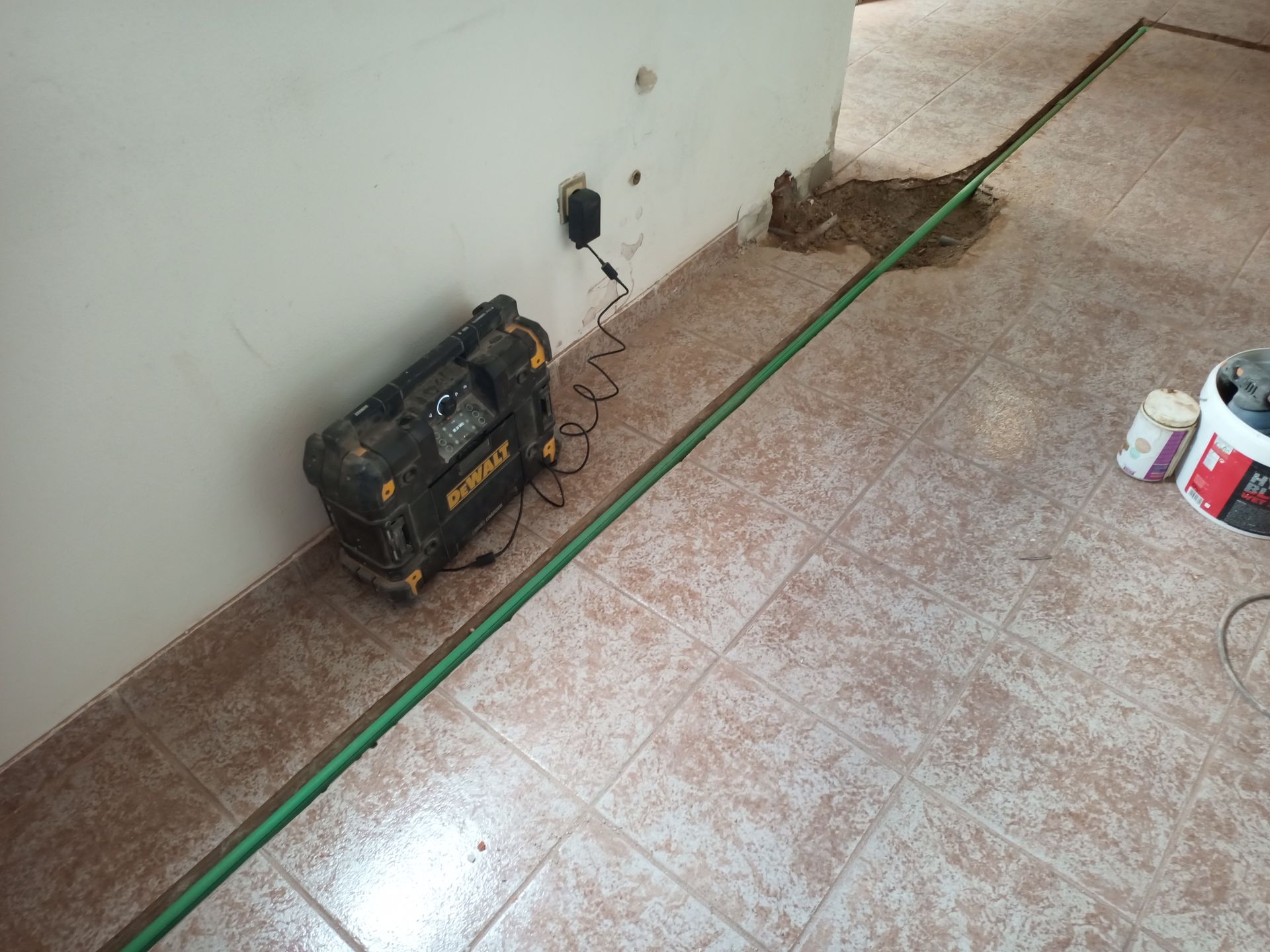 Caixa de ferramenta elétrica preta e amarela carregando, ao lado de tubos verdes no piso de azulejos perto da parede.