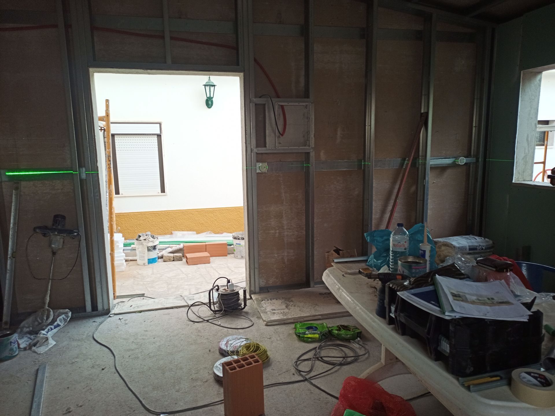 Interior de uma sala em construção, com porta aberta para um pátio externo, ferramentas e materiais espalhados.