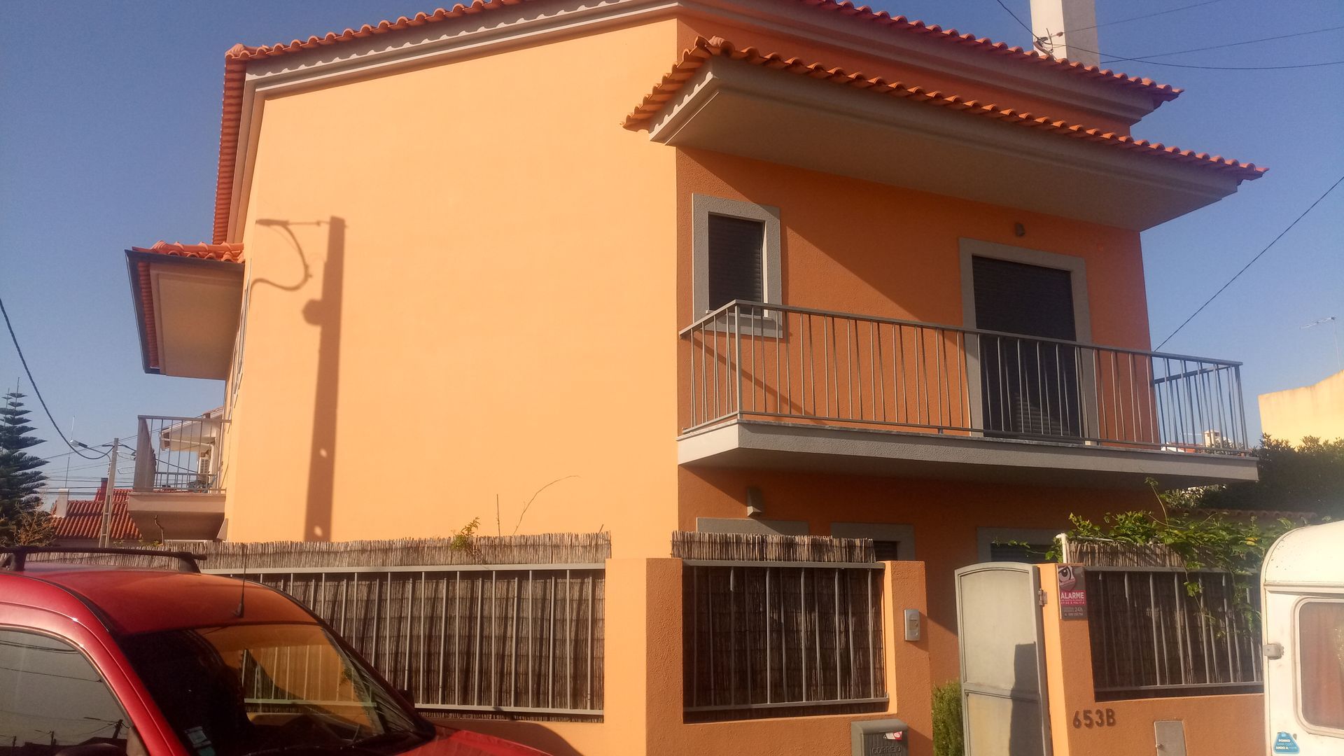 Casa laranja de dois andares com uma pequena varanda, telhado marrom e cerca de madeira na frente. Um carro vermelho é parcialmente visível.