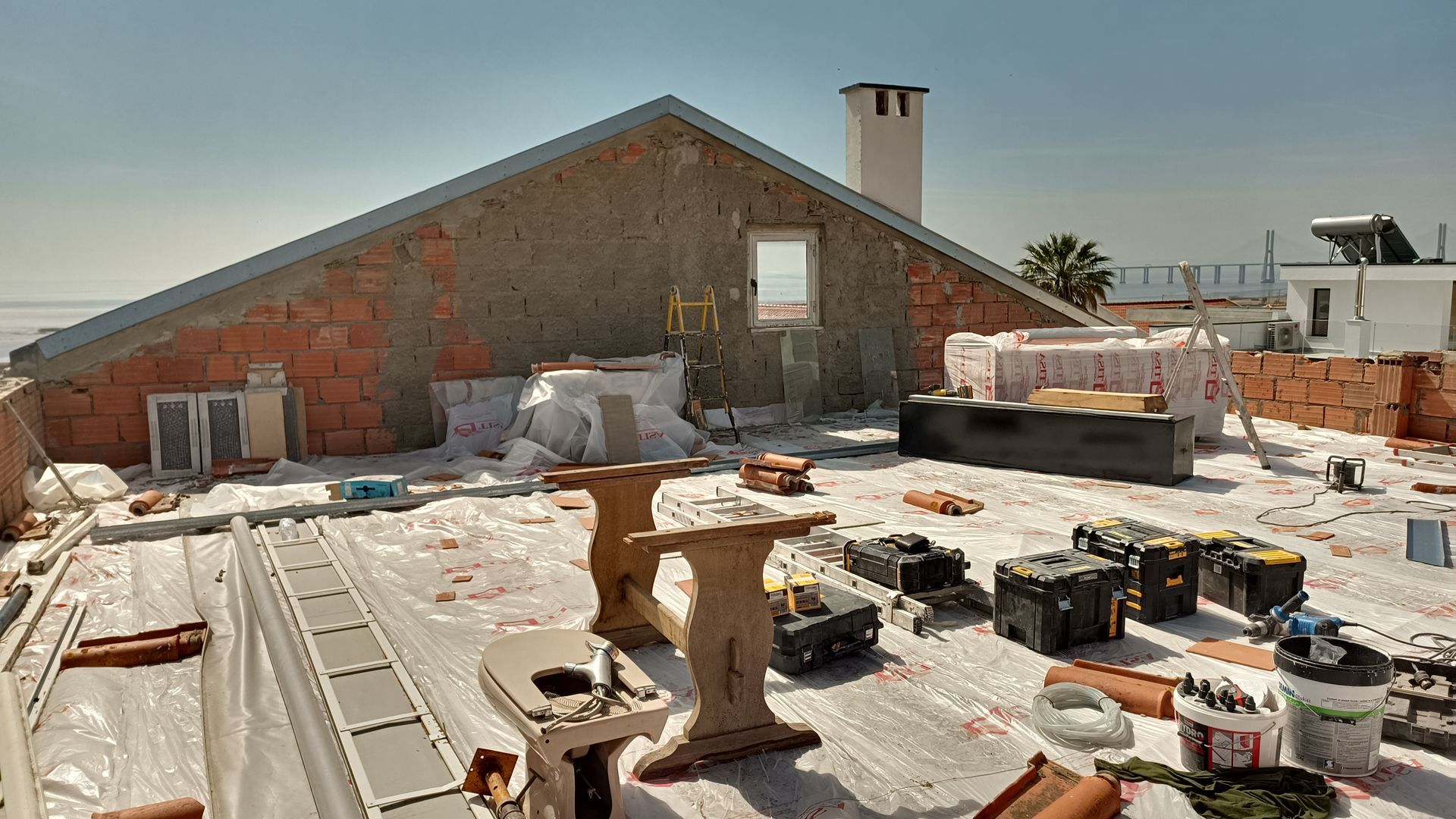 Obra em telhado. Parede de tijolos vermelhos e bege sob céu azul. Ferramentas, materiais e uma chaminé são visíveis.