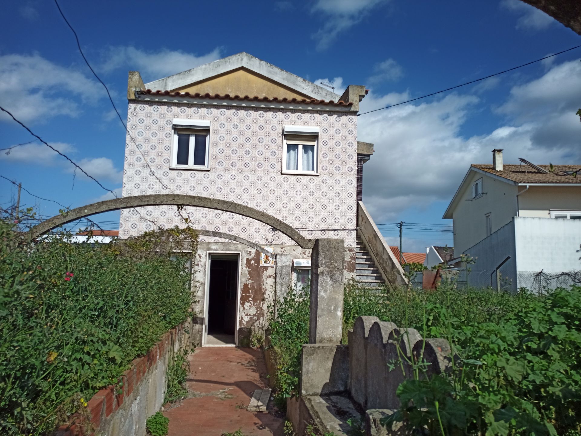 Prédio de dois andares com fachada estampada, porta aberta, entrada em arco, escadas e plantas, sob um céu azul.