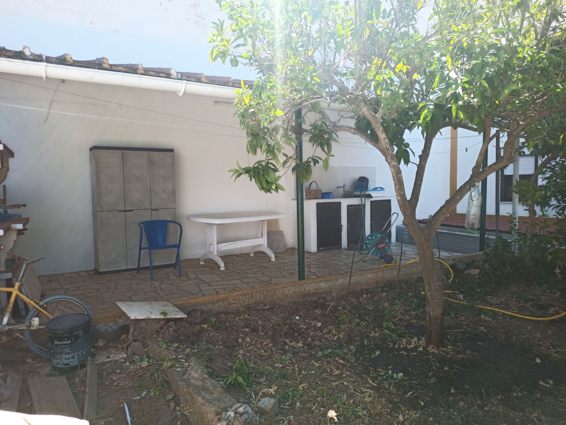 Quintal com parede branca, uma árvore, uma mesa e outros objetos de exterior. A luz do sol incide sobre ele.