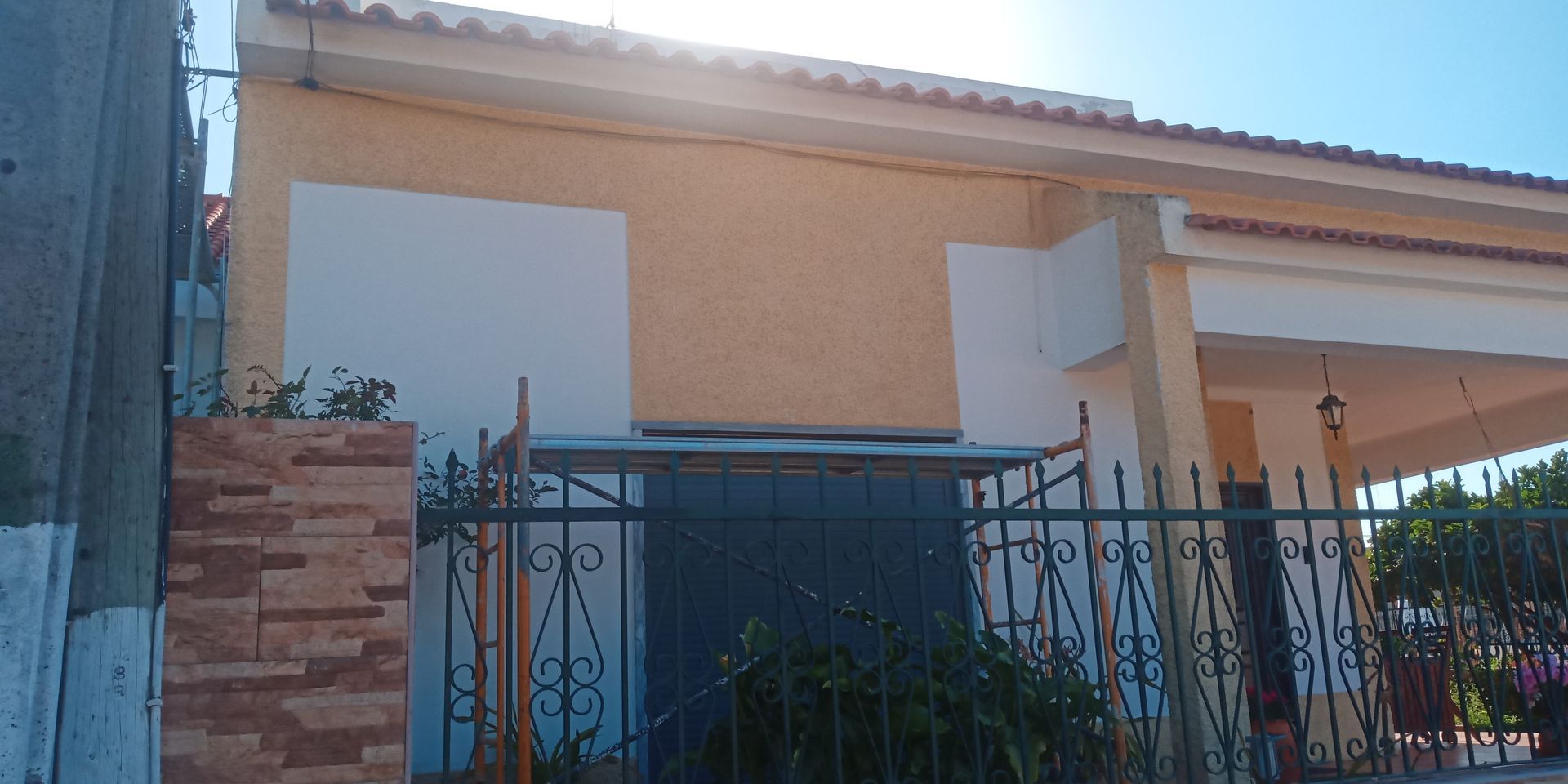 Um prédio amarelo com um portão de metal escuro e um muro de tijolos na frente. Céu azul.