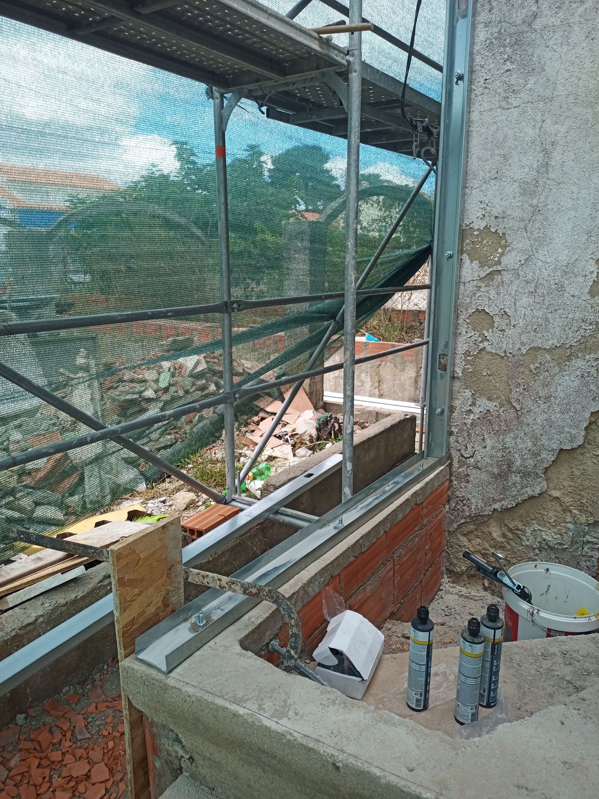 Janela em construção com andaimes e redes de segurança, alvenaria e concreto.