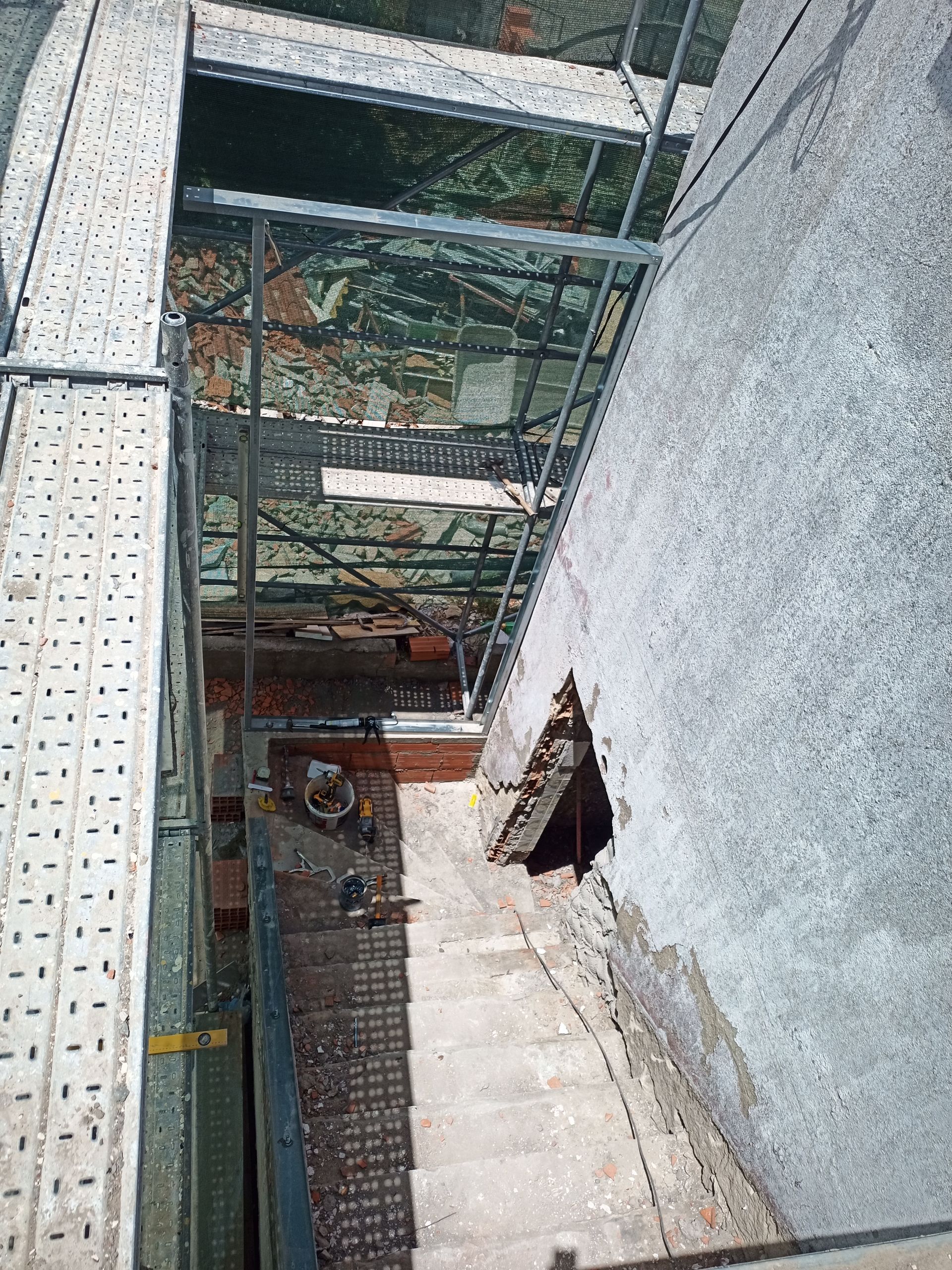 Olhando para cima, avista-se uma estrutura estreita e deteriorada: paredes de concreto, andaimes metálicos, alvenaria e um buraco na parede.
