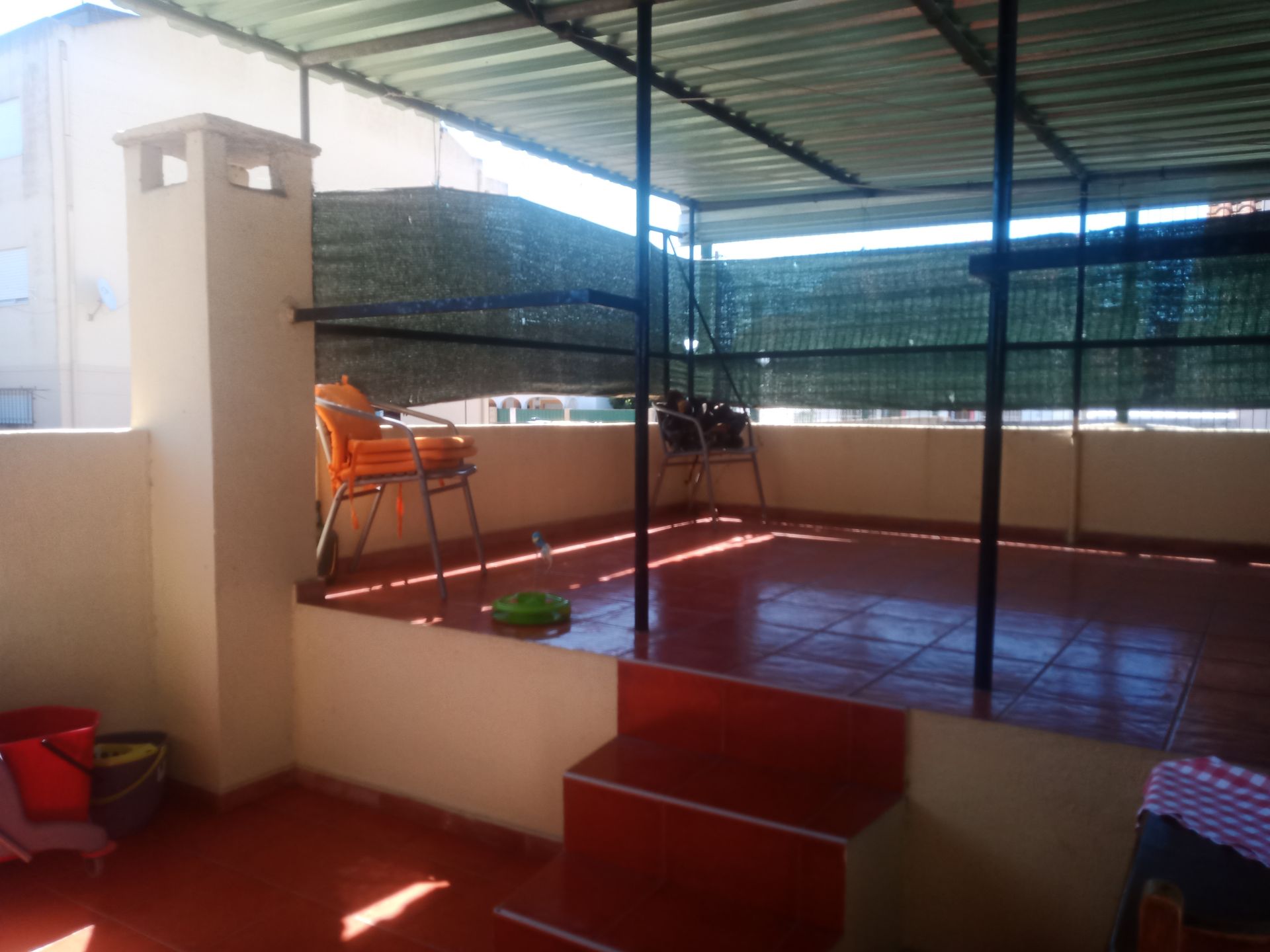 Terraço na cobertura com piso de azulejo, sombreado por um telhado de zinco. Duas cadeiras laranjas. Degrau e chaminé vermelhos.