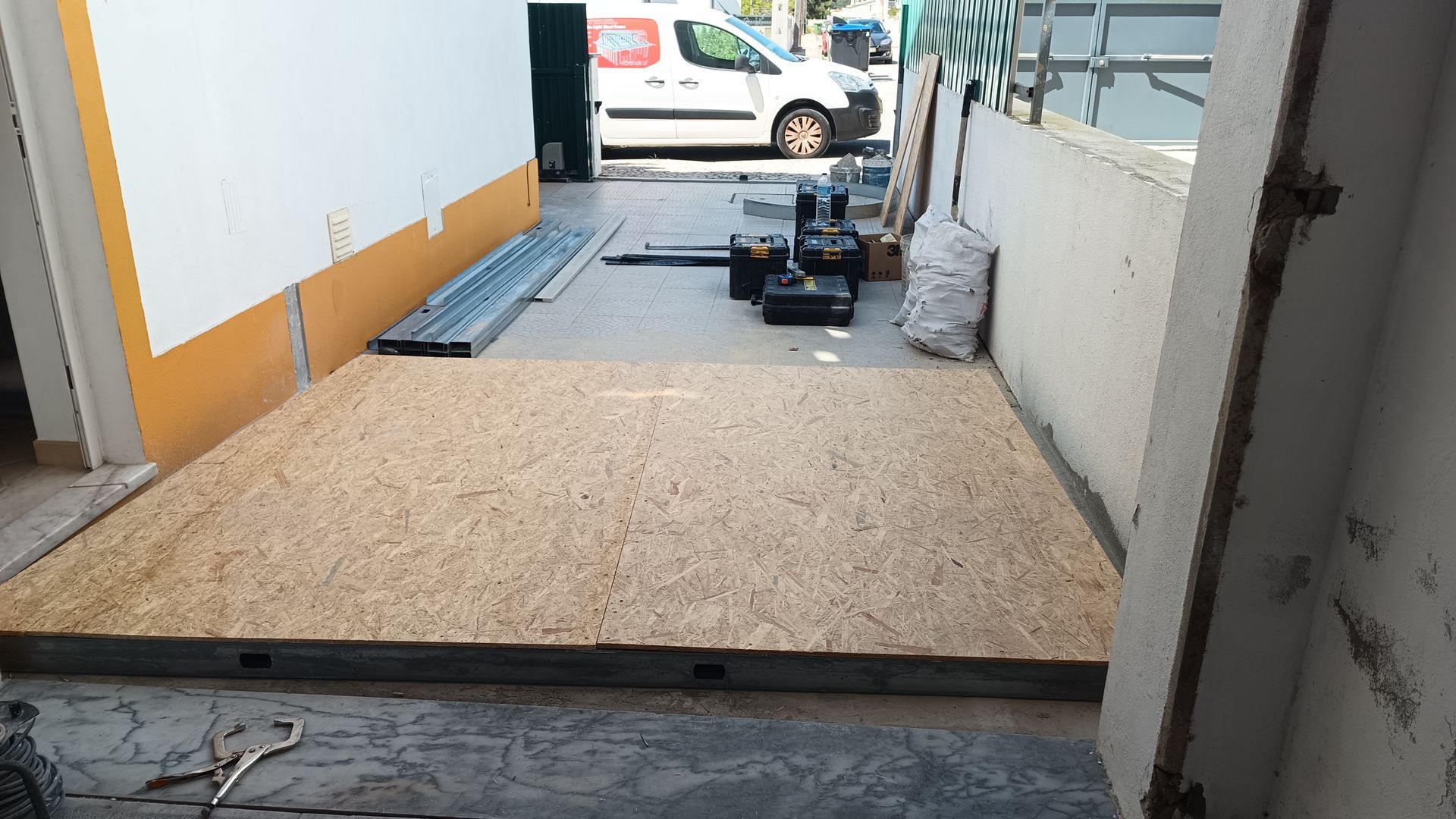 Grande painel de madeira no chão, com ferramentas e uma van ao fundo, visto de uma porta.