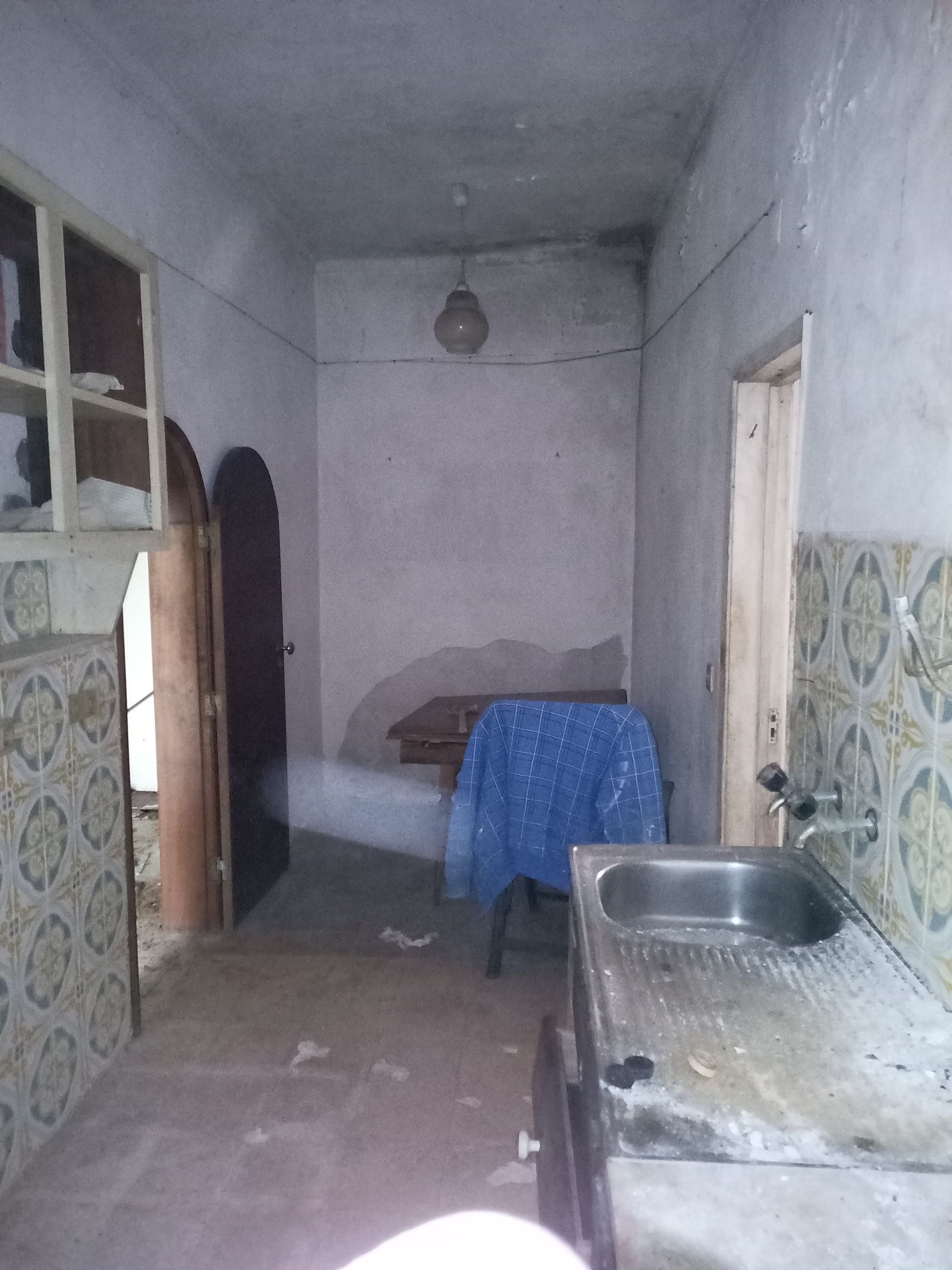 Cozinha estreita com pia, armário e porta. Paredes lisas, superfícies desgastadas, iluminação fraca.