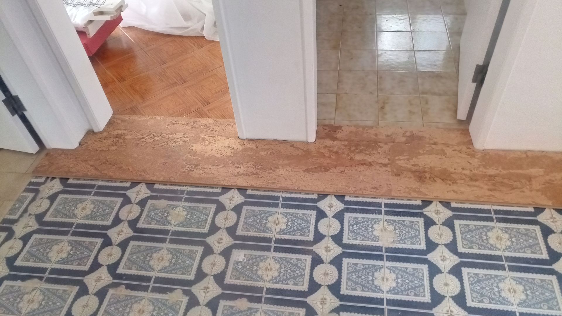 Transição de piso: ladrilho estampado, faixa de concreto, madeira e ladrilho bege conduzem a três portas.