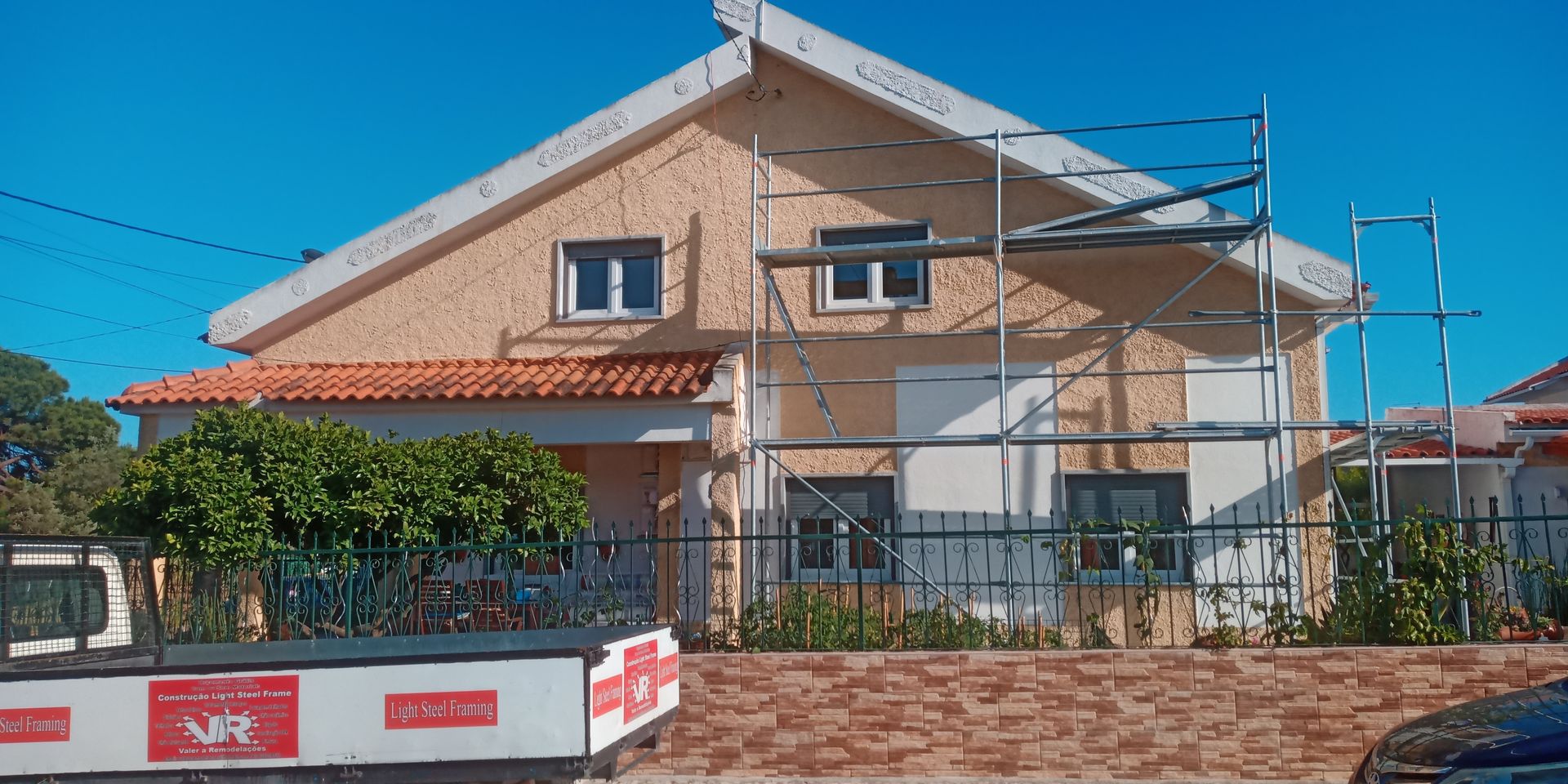 Casa bege de dois andares com andaimes, sob um céu azul claro. Muro de tijolos e arbustos na frente.