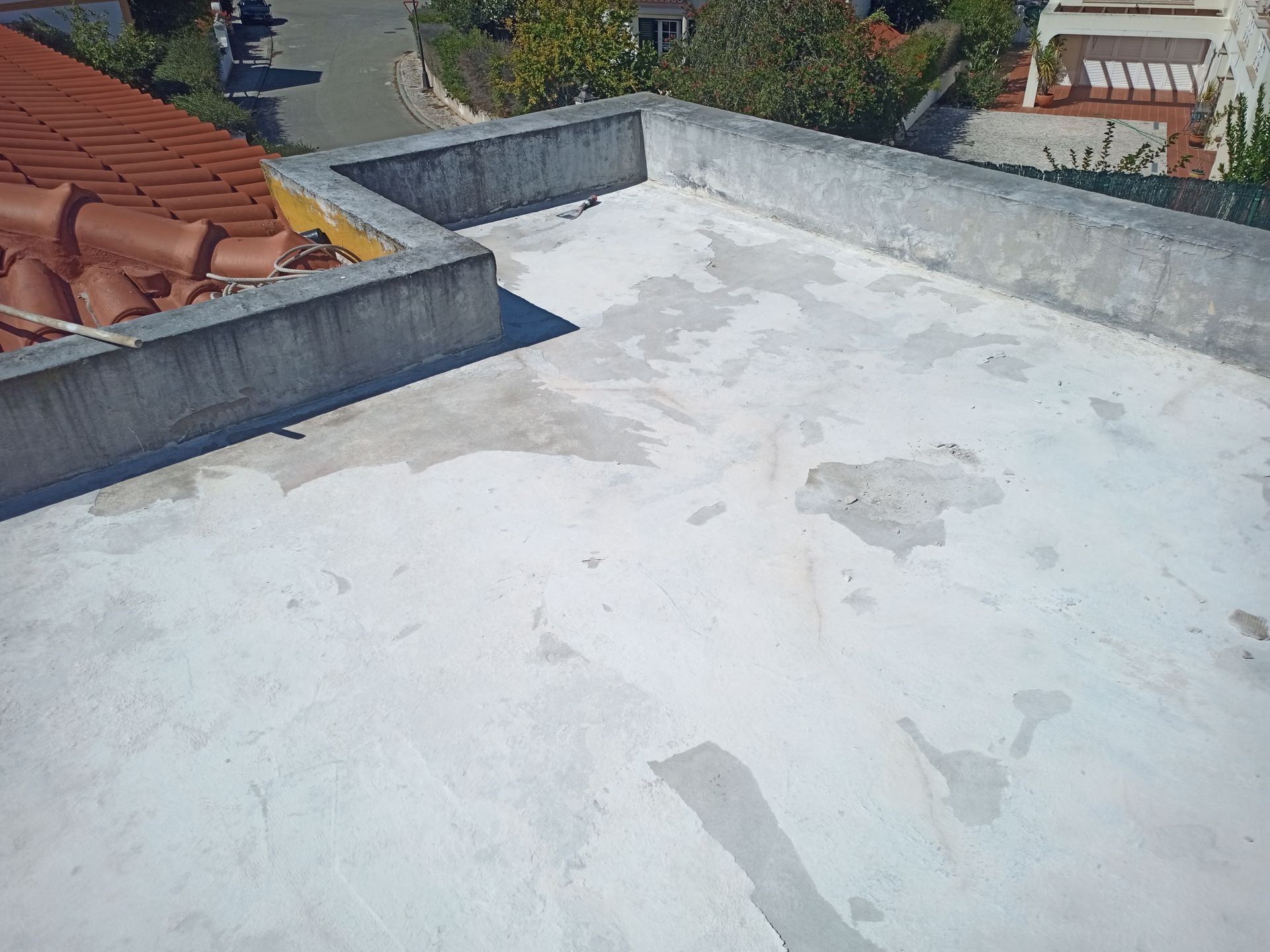 Telhado plano branco com perímetro de concreto, remendos visíveis e telha vermelha ao fundo.