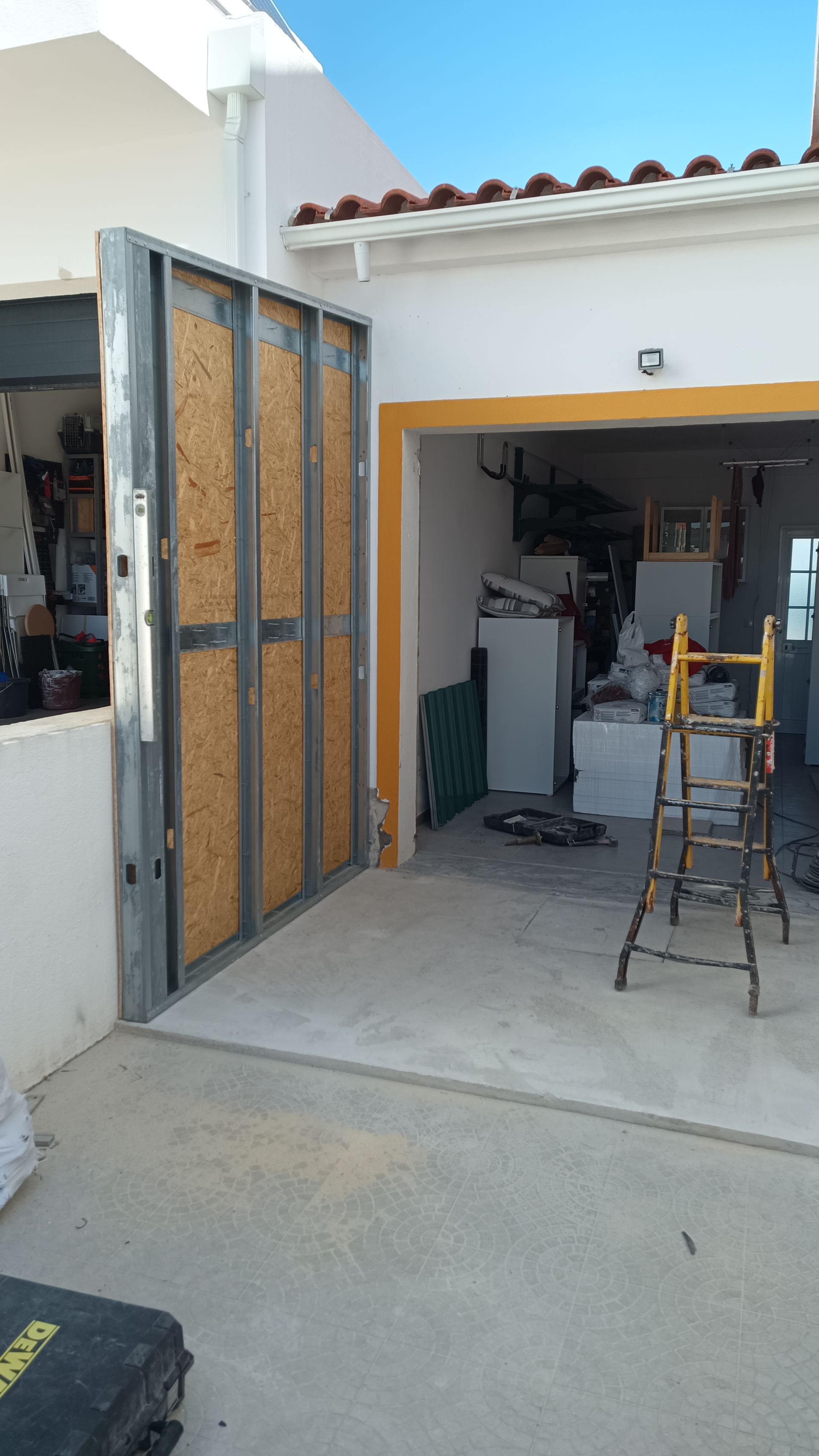 Vista externa de uma garagem em construção. Estrutura metálica com painéis de madeira, ferramentas e materiais visíveis. Dia ensolarado.