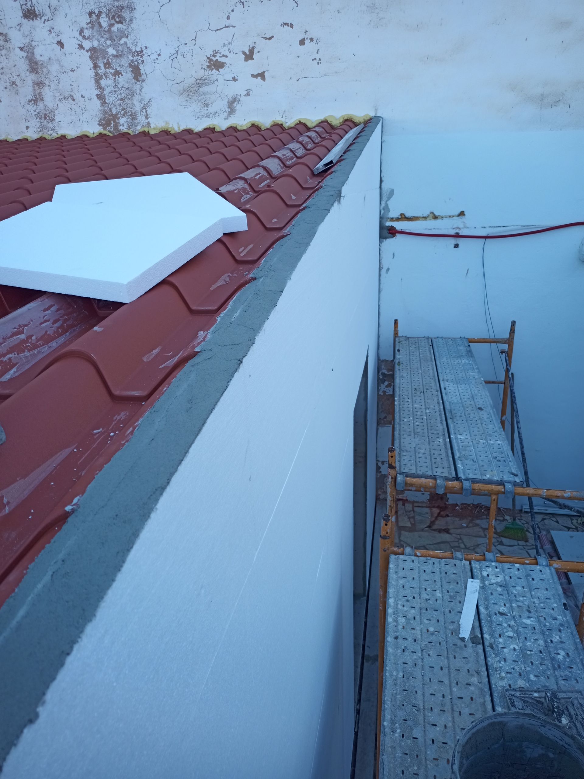 Borda do telhado de telhas vermelhas ao lado de uma parede branca em construção; andaimes abaixo; acabamento cinza.