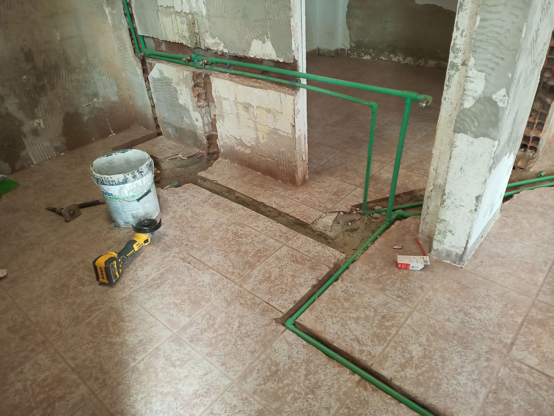 Tubos verdes dispostos sobre piso de azulejo, canteiro de obras visível. Balde branco, ferramentas e paredes parcialmente acabadas.