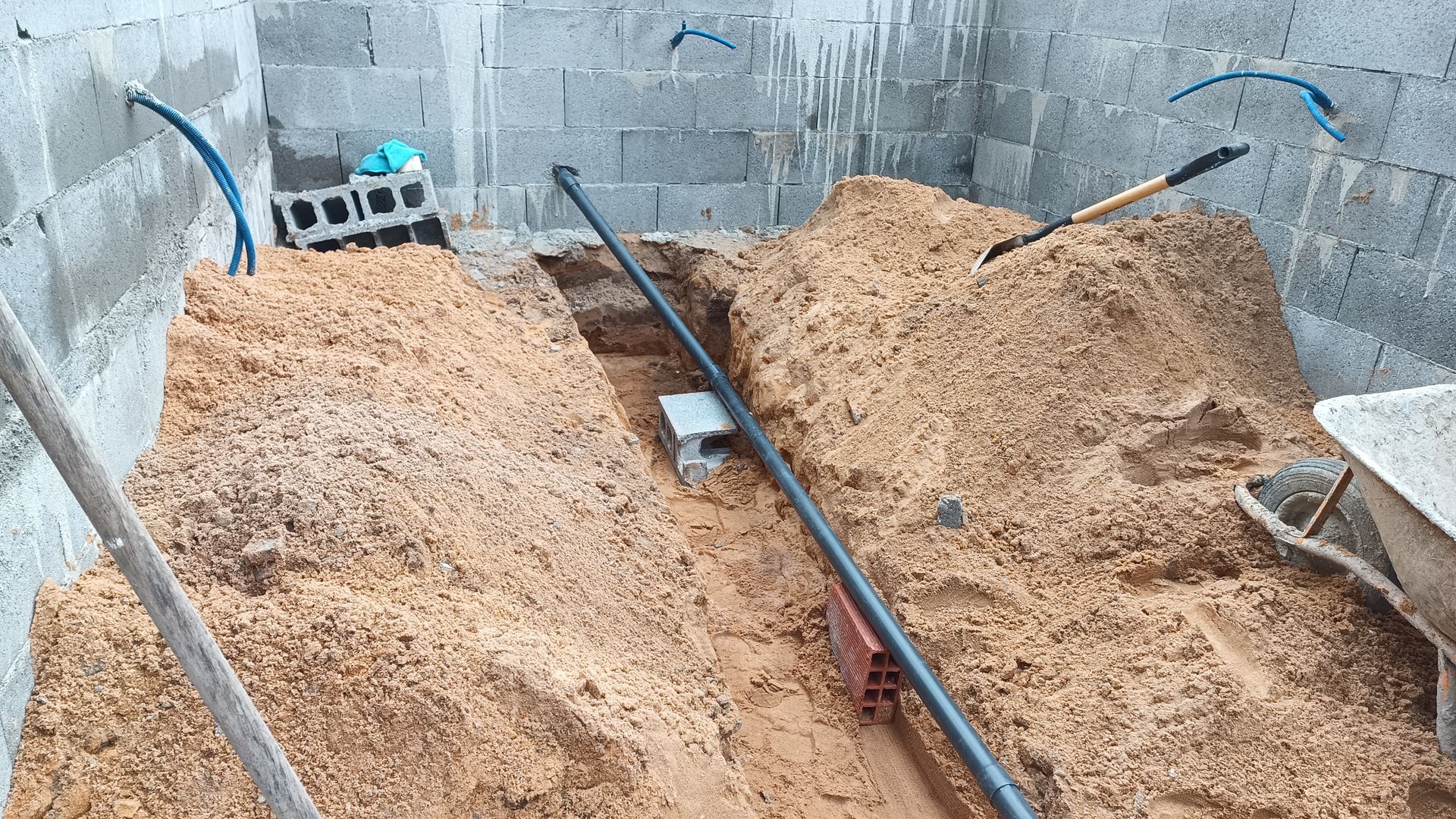 Canteiro de obras: Vala com cano preto, areia, paredes de tijolo e ferramentas.