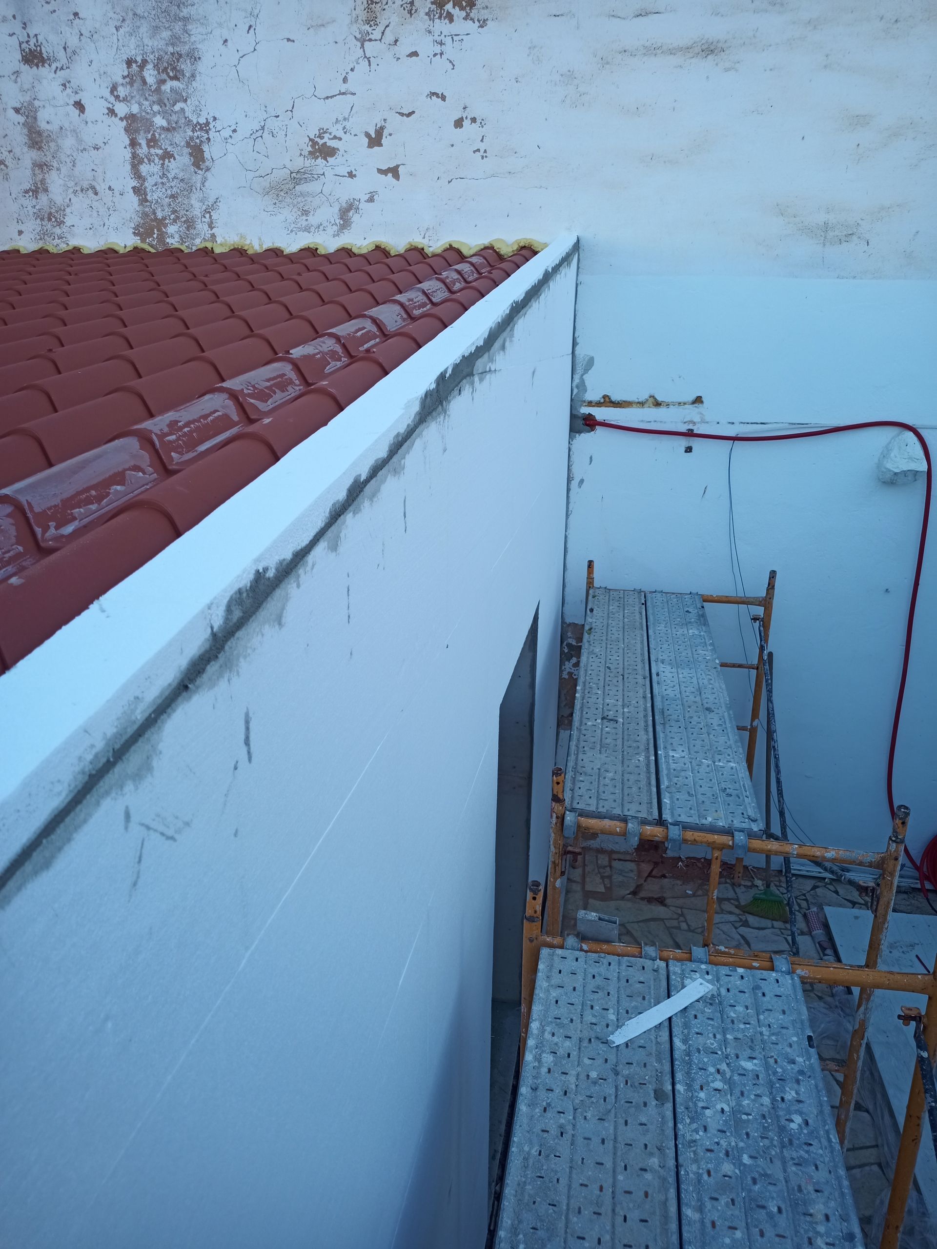 Telhado com telhas vermelhas encontra parede branca, andaimes no canto.