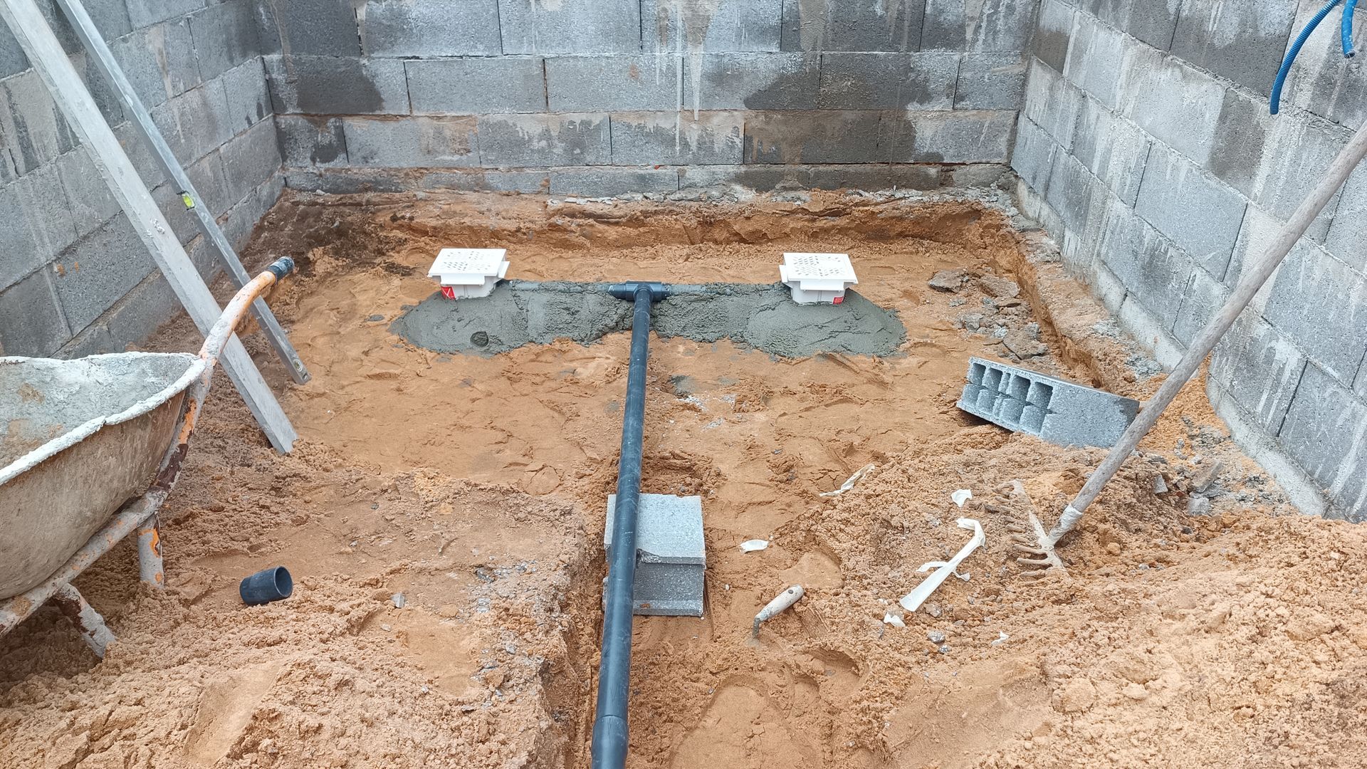 Canteiro de obras com instalações hidráulicas; canos pretos em solo arenoso, blocos de cimento, carrinho de mão e paredes de blocos de concreto cinza.