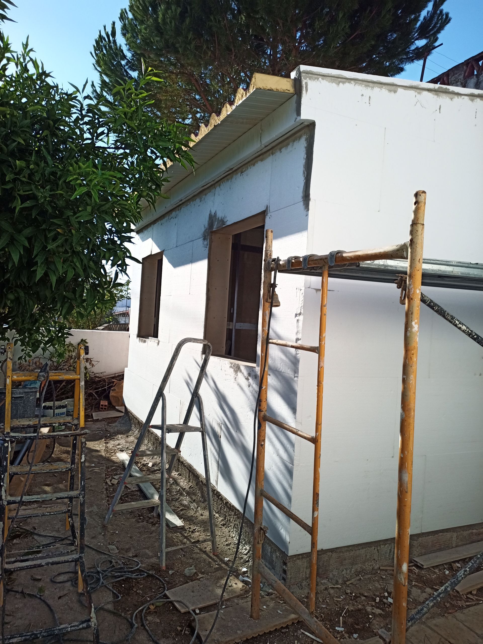 Vista exterior de um edifício branco em construção, com andaimes e duas janelas; uma árvore está à esquerda.