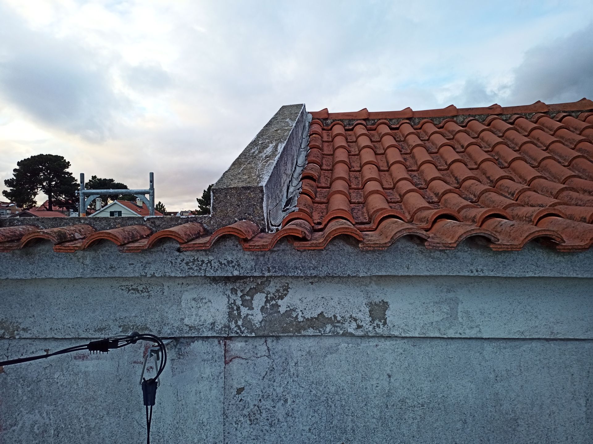 Borda do telhado de telha vermelha, parede cinza, céu nublado.