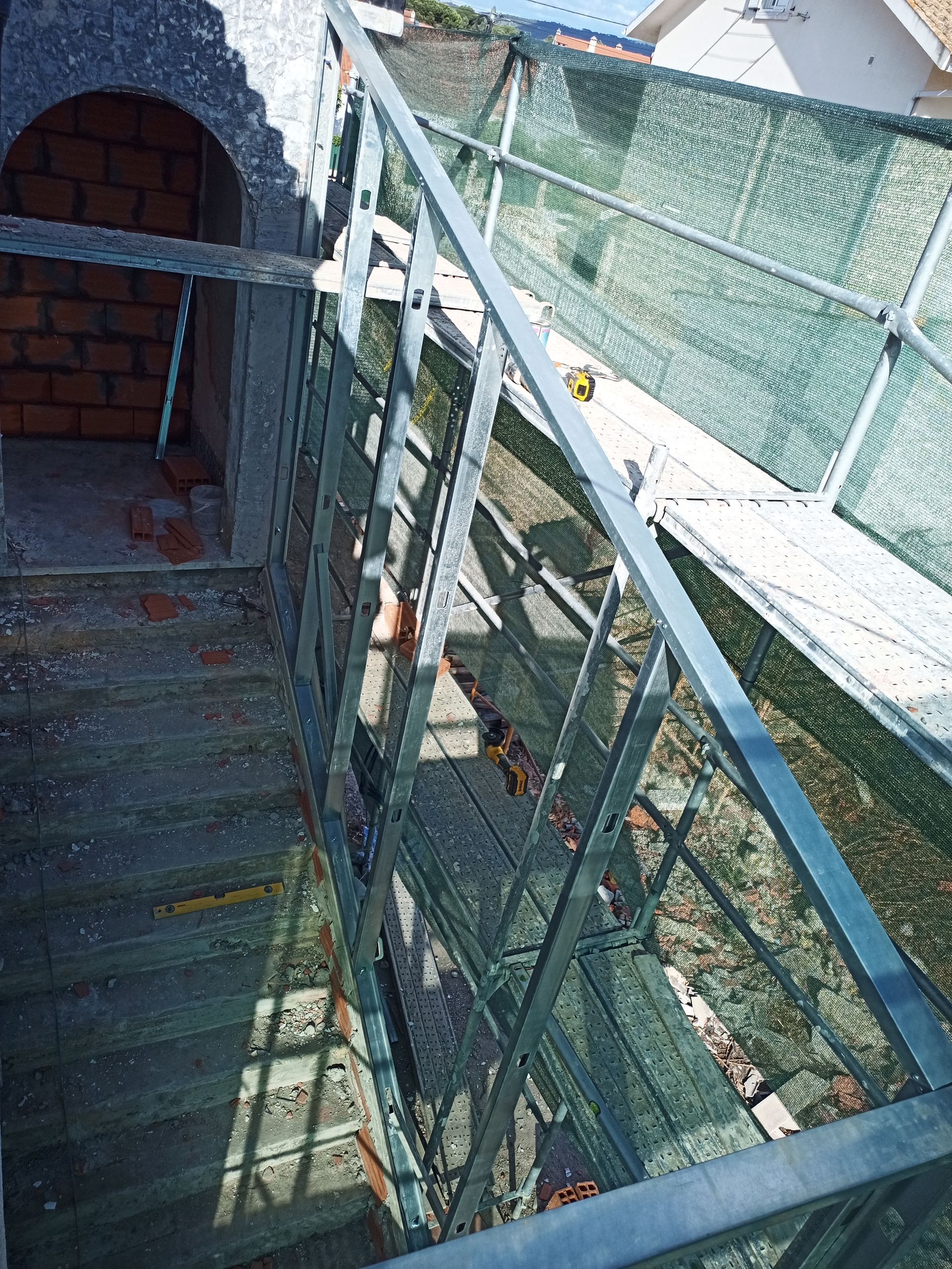 Escada com corrimão de metal e rede verde, vista de cima. Parede de tijolos, luz solar.