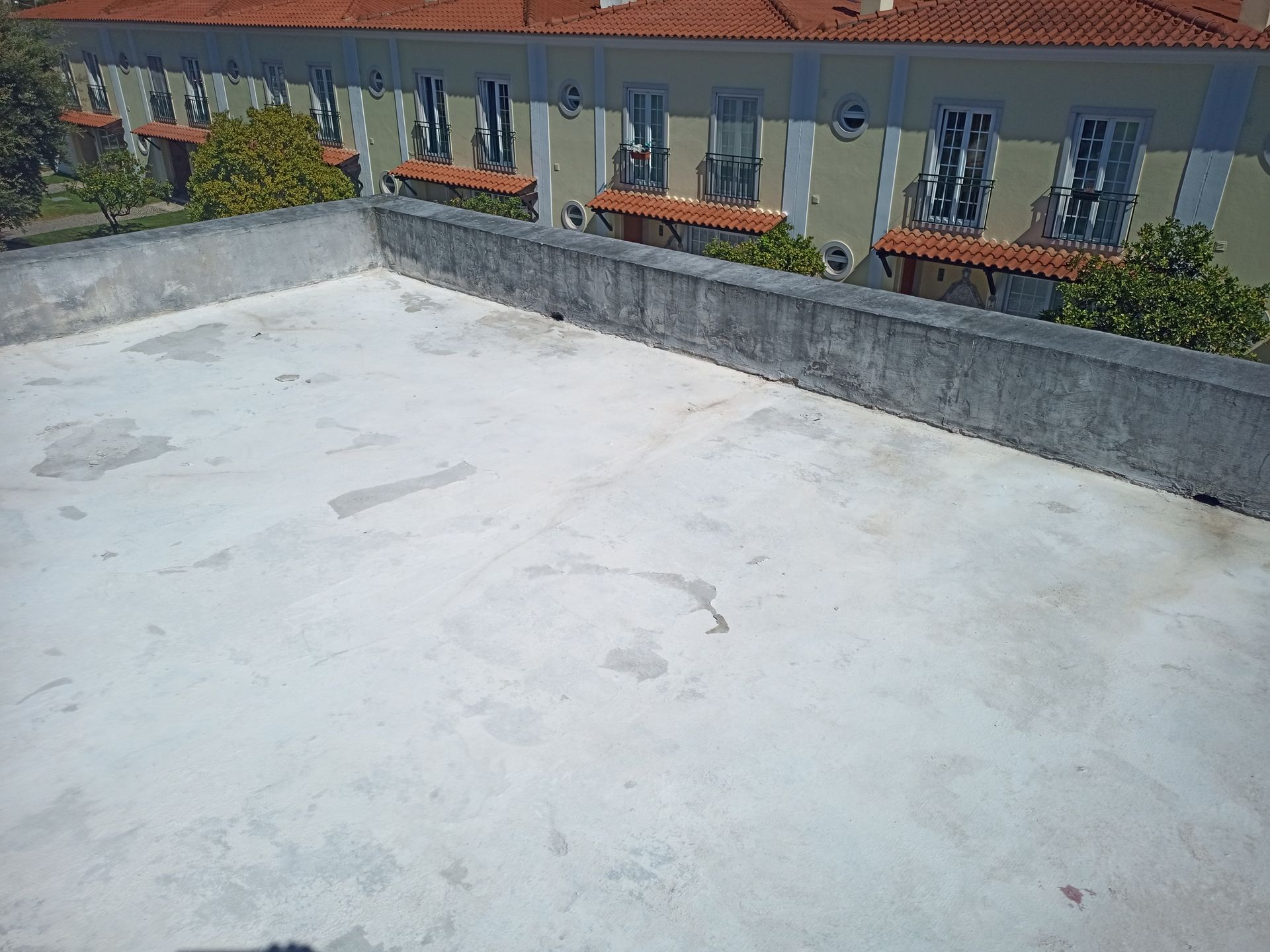 Telhado plano e claro com uma pequena barreira de concreto. Prédio com janelas e telhado de telha vermelha ao fundo.