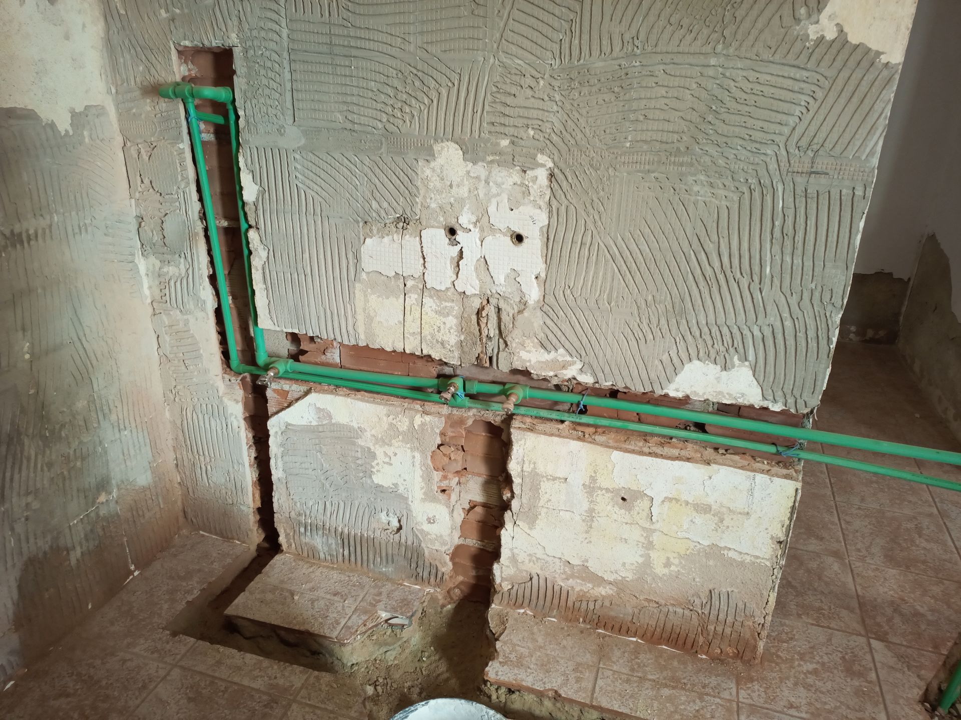 Reforma de um banheiro, com encanamento exposto contra uma parede de tijolos e azulejos danificada, canos verdes visíveis.
