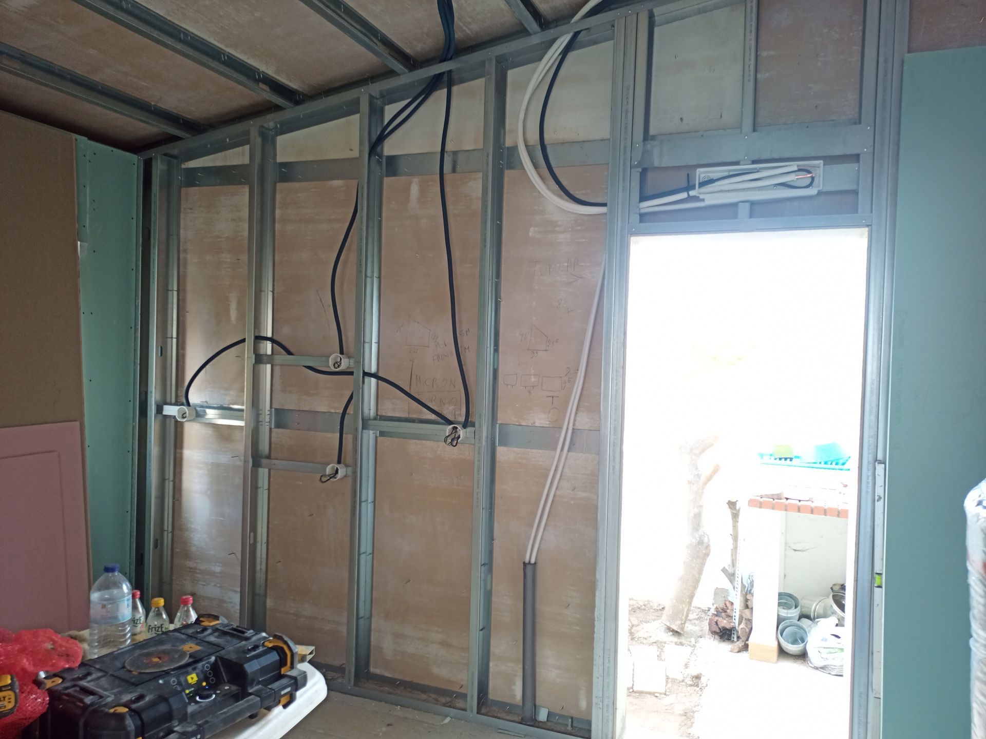 Interior de um cômodo em construção com estrutura metálica, compensado, fiação elétrica e uma porta.