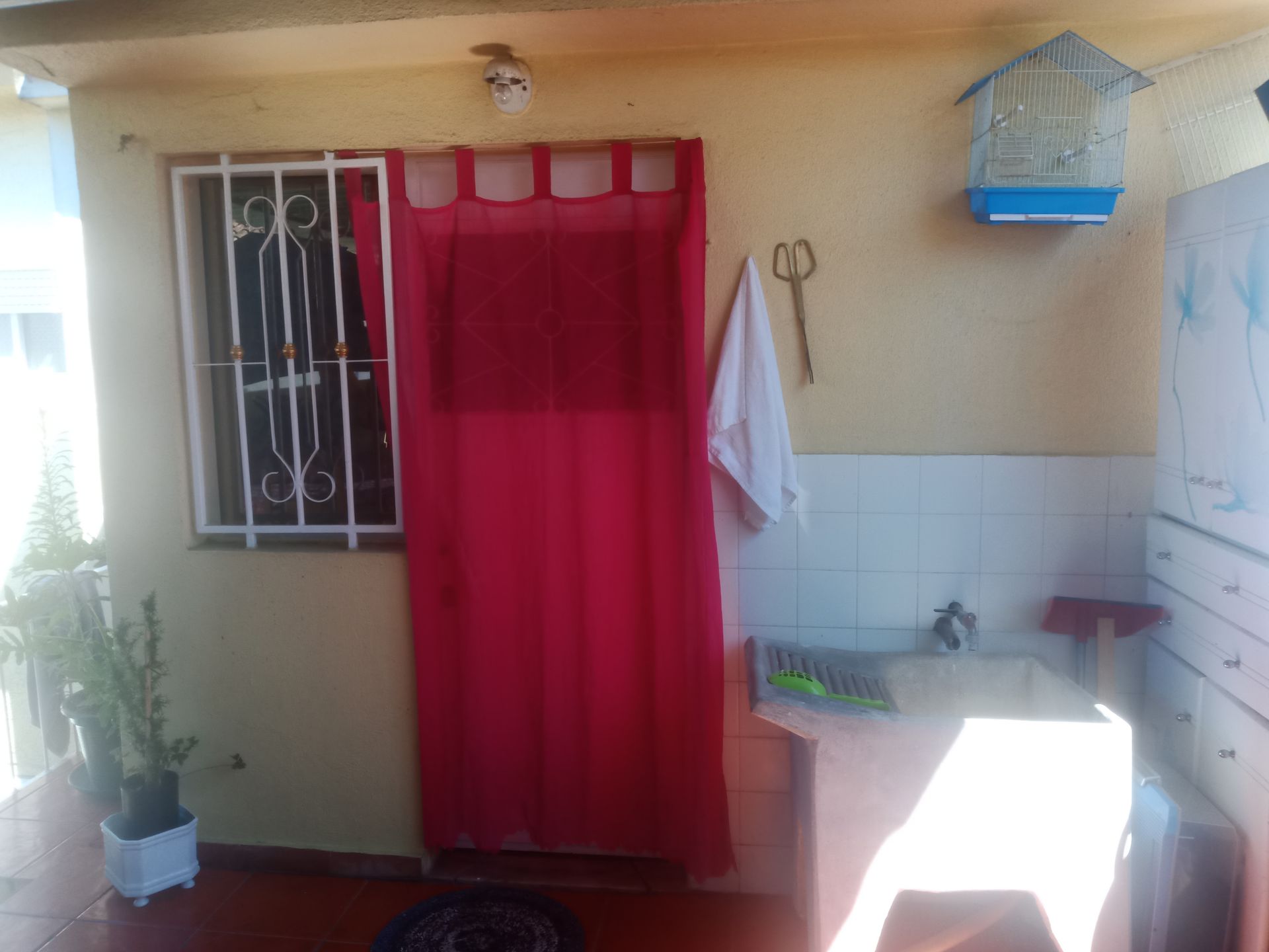Pequena cozinha externa com cortina vermelha sobre a porta, janela com grades e gaiola para pássaros.