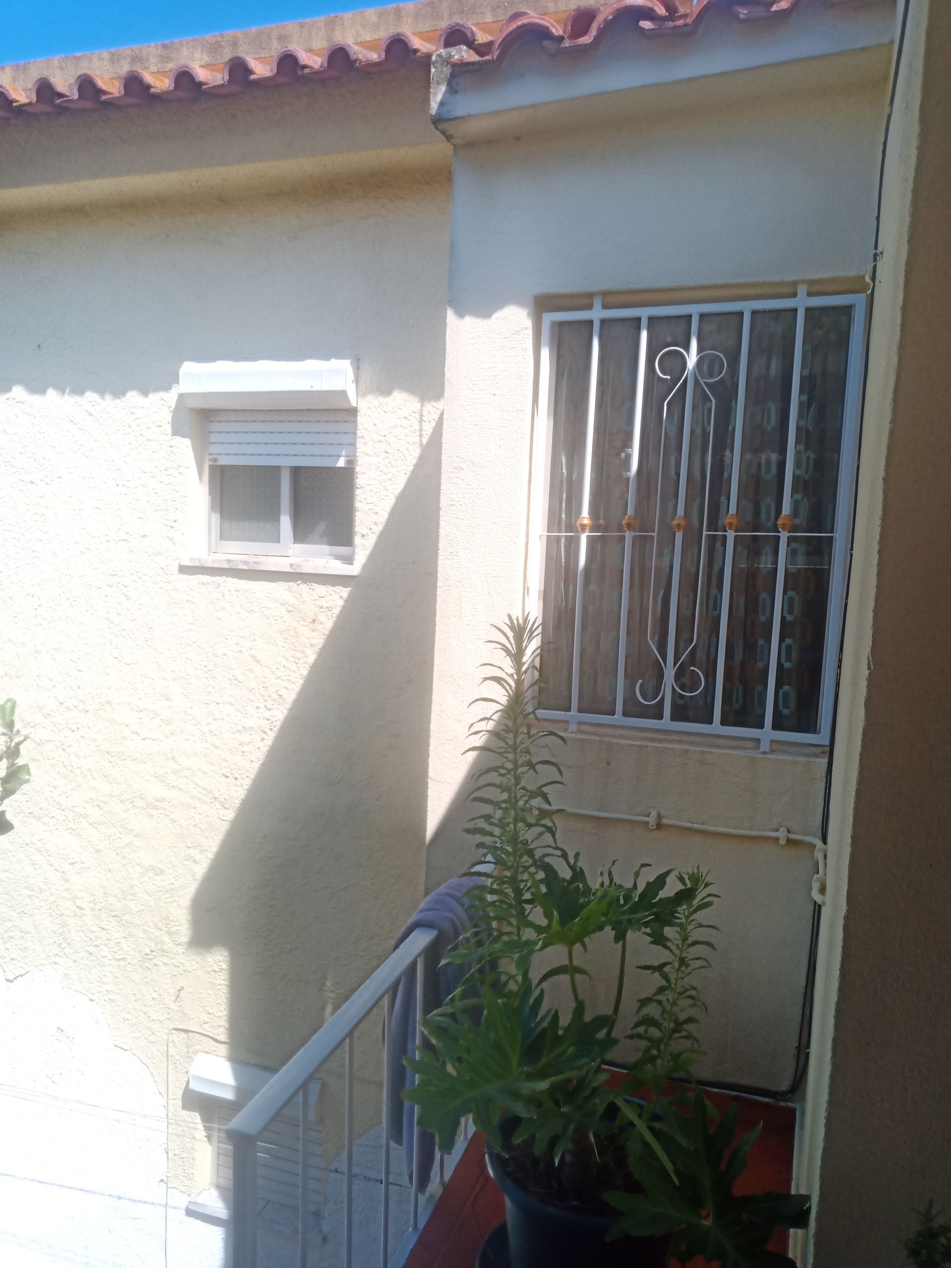 Vista exterior de um edifício com duas janelas. Uma janela tem uma persiana de enrolar; a outra tem grades de segurança.