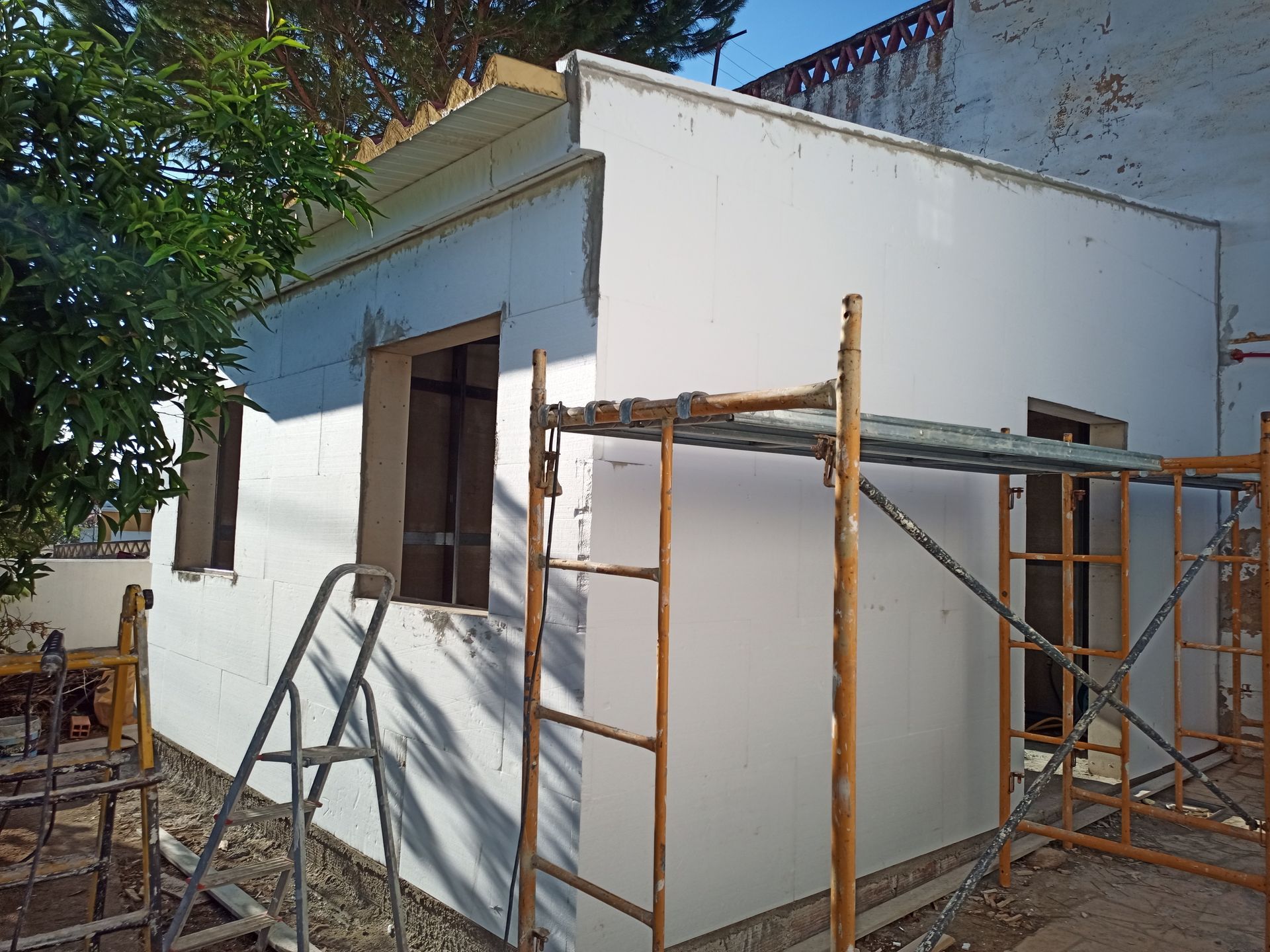 Vista exterior de um edifício em construção, com paredes brancas, andaimes e árvores ao fundo.