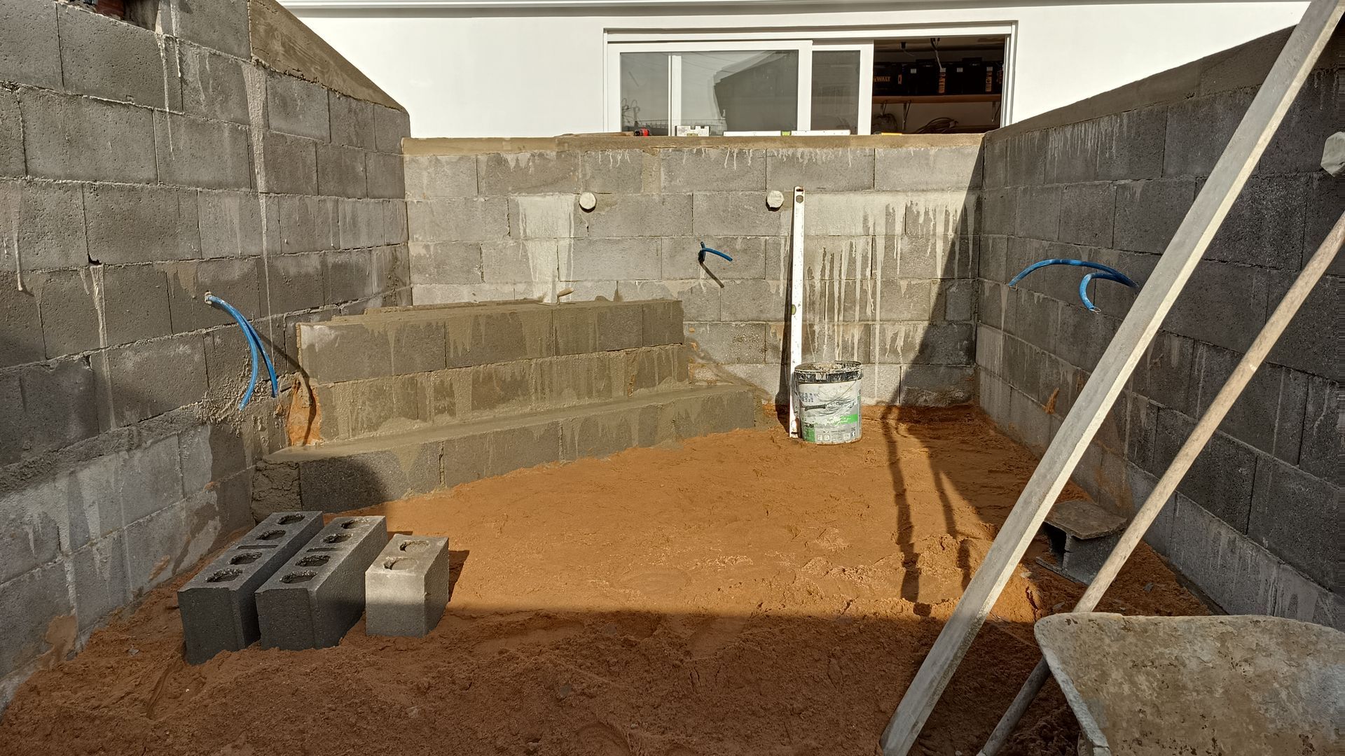 Canteiro de obras: estrutura de concreto inacabada com degraus, blocos expostos, piso de terra e um prédio branco parcialmente visível ao fundo.