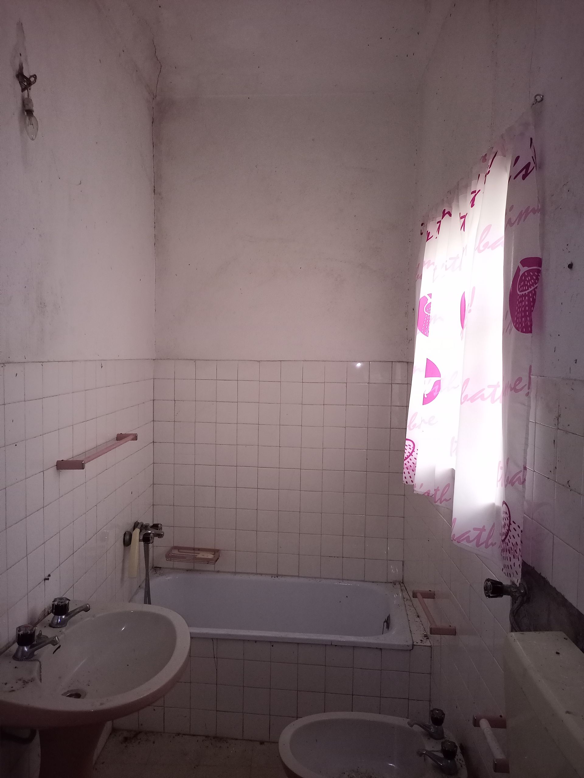 Um pequeno banheiro com azulejos brancos, banheira, pia, bidê e uma janela com cortina rosa estampada.