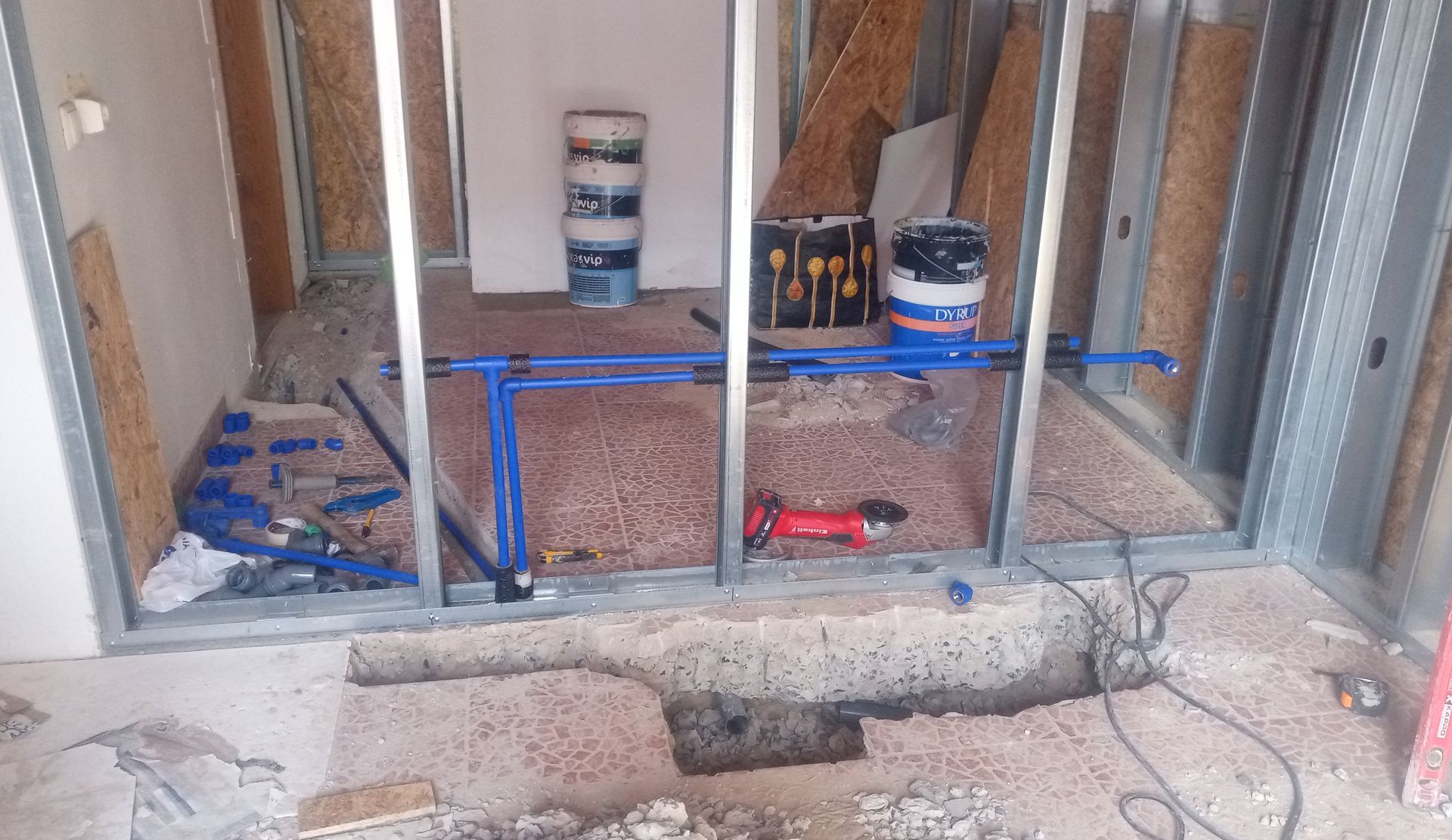 Construção de um banheiro, com estrutura metálica nas paredes, canos azuis, piso de concreto, latas de tinta e ferramentas visíveis.