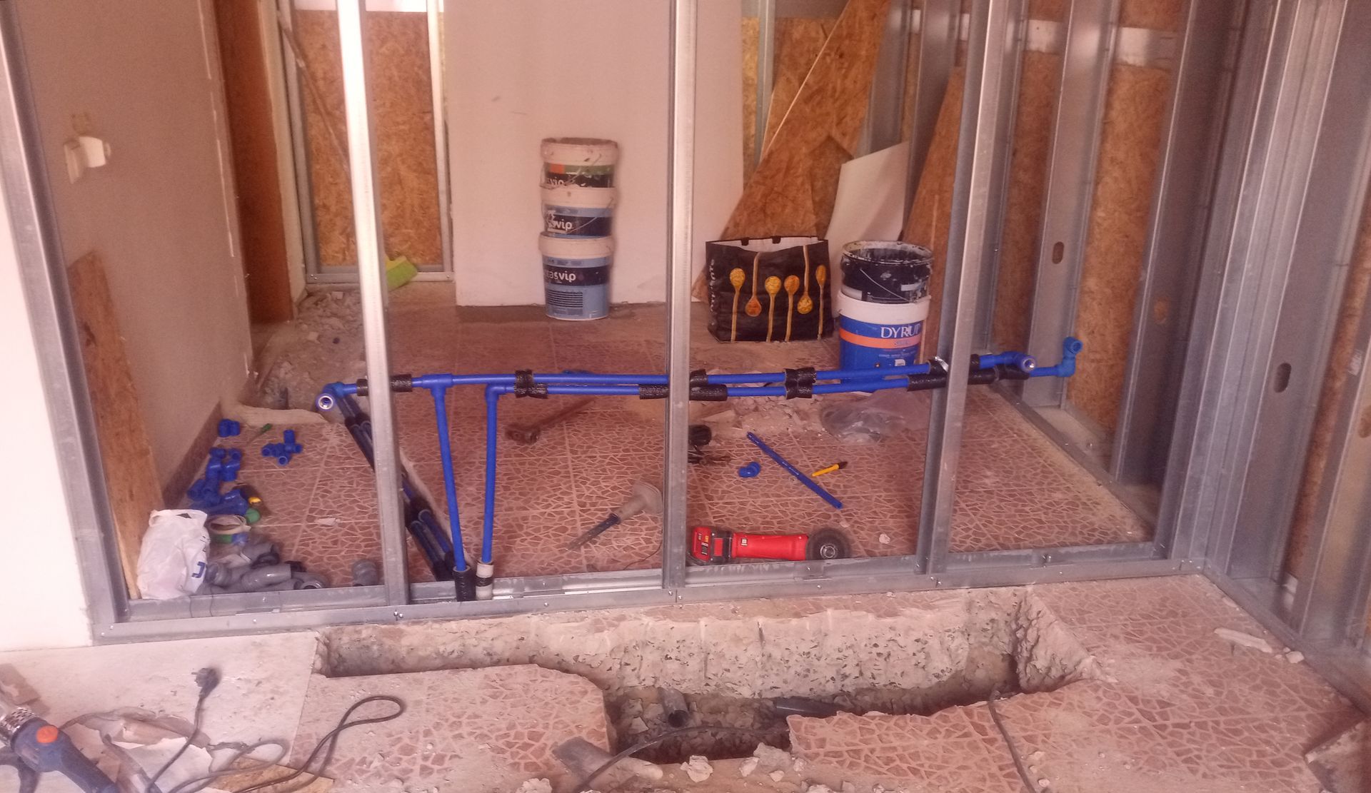 Interior de um canteiro de obras com estrutura metálica, encanamento azul e entulho no chão.