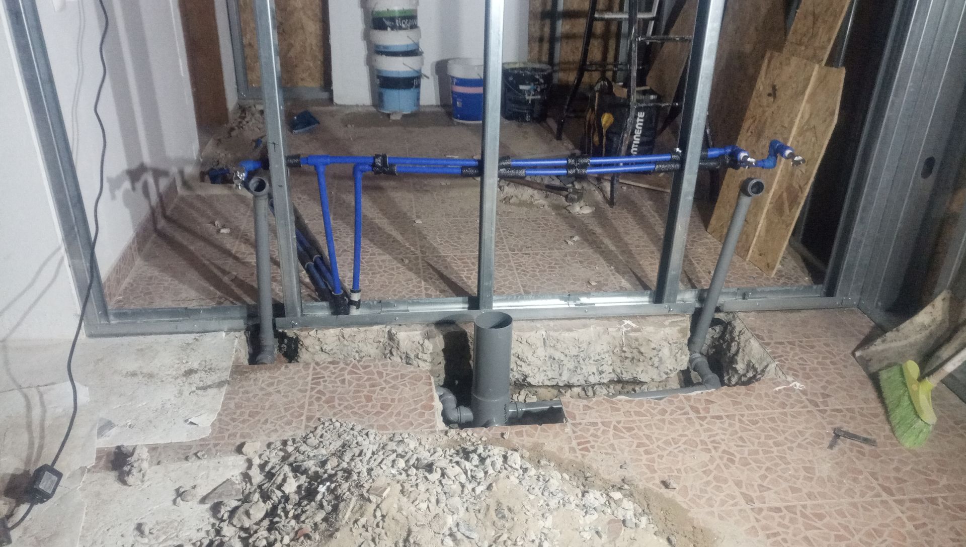 Canteiro de obras: Encanamento exposto, estrutura metálica, piso de azulejo quebrado e entulho.