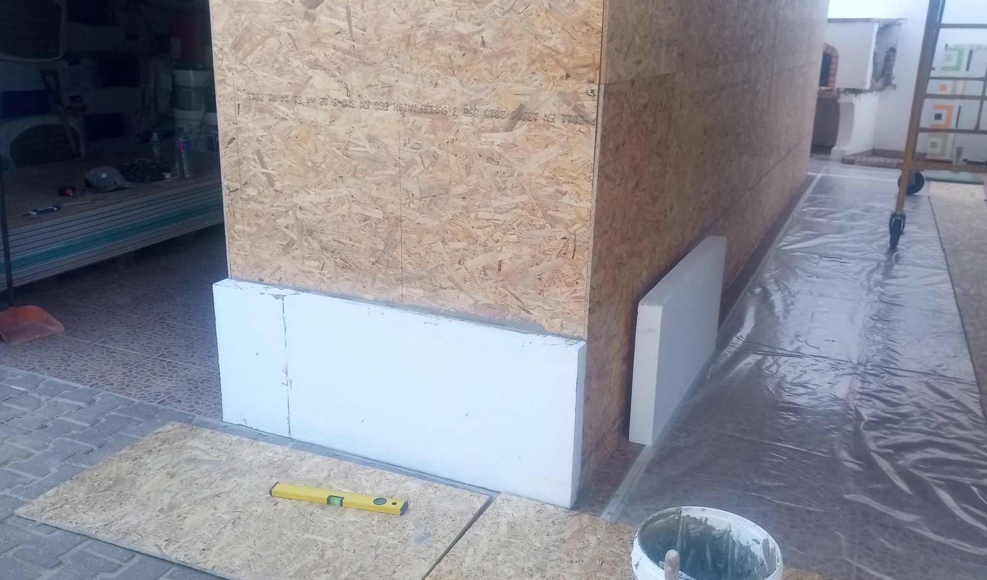 Parede de OSB com isolamento branco sendo instalada na base. Um nível e um balde de material são visíveis.