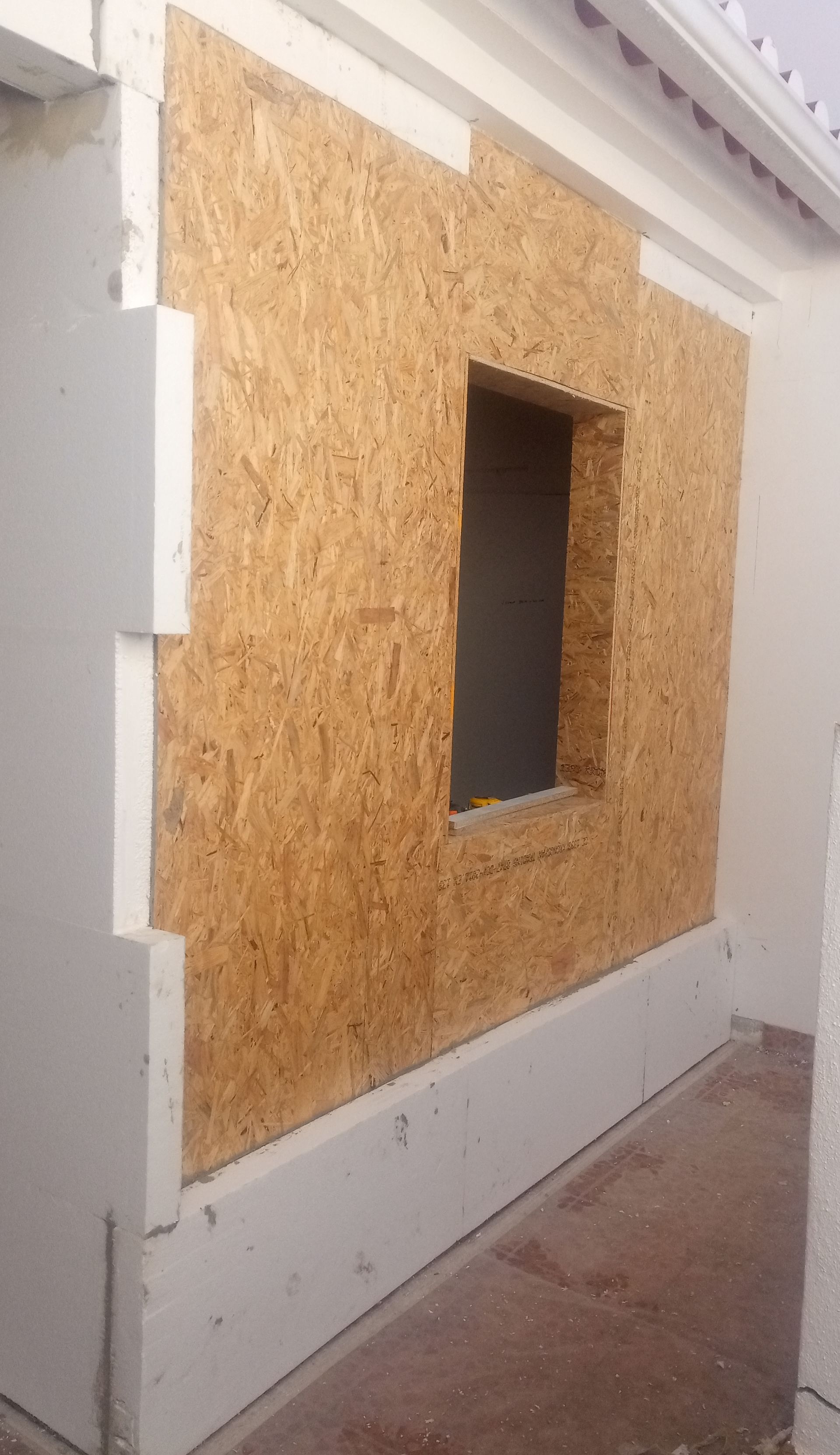 Parede externa em construção com OSB e abertura para janela. Acabamento e rodapé brancos.