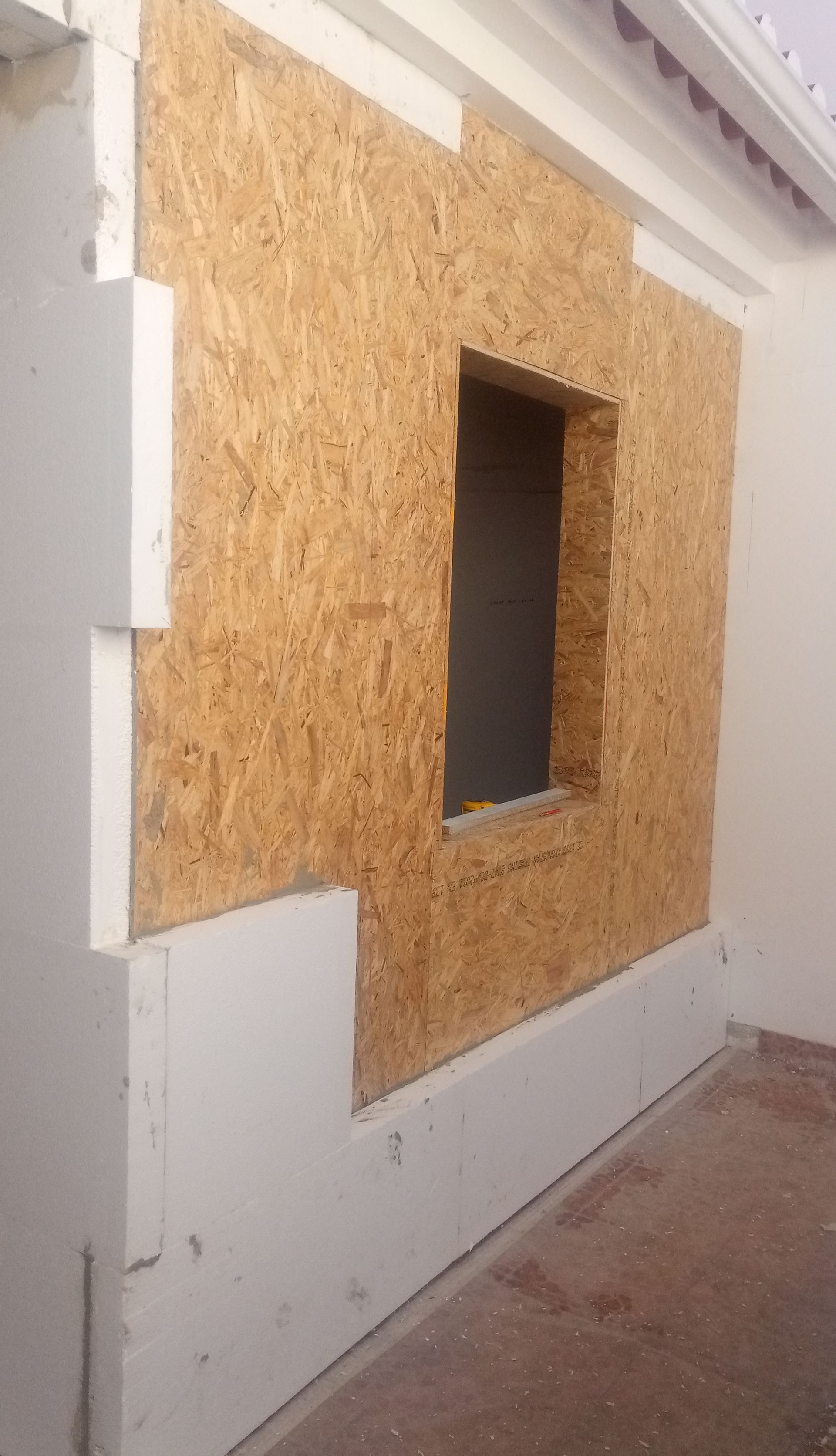 Parede externa em construção, mostrando painéis de OSB expostos ao redor de uma janela e isolamento em camadas.