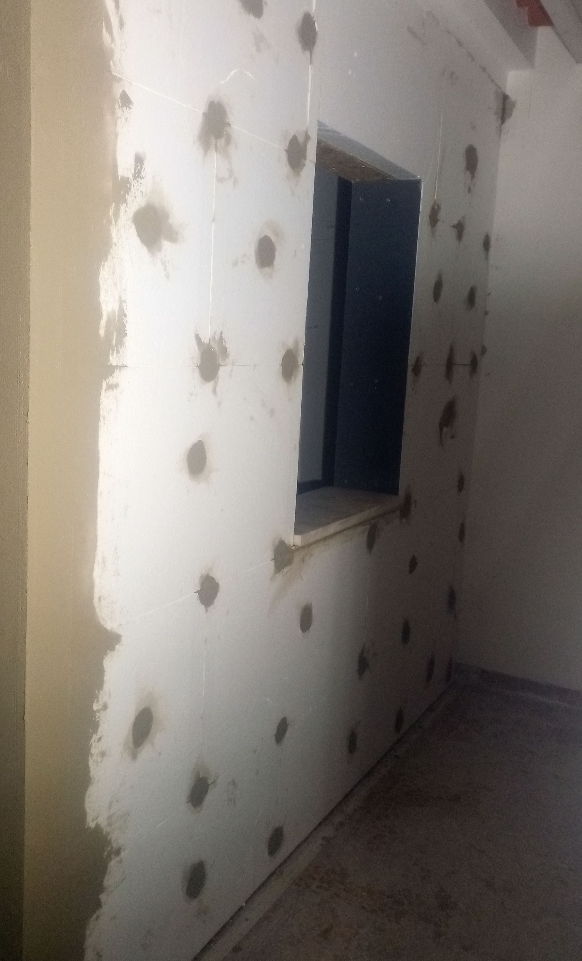 Parede interna com diversas manchas cinzentas, possivelmente para fixação de isolamento, ao redor de uma janela.