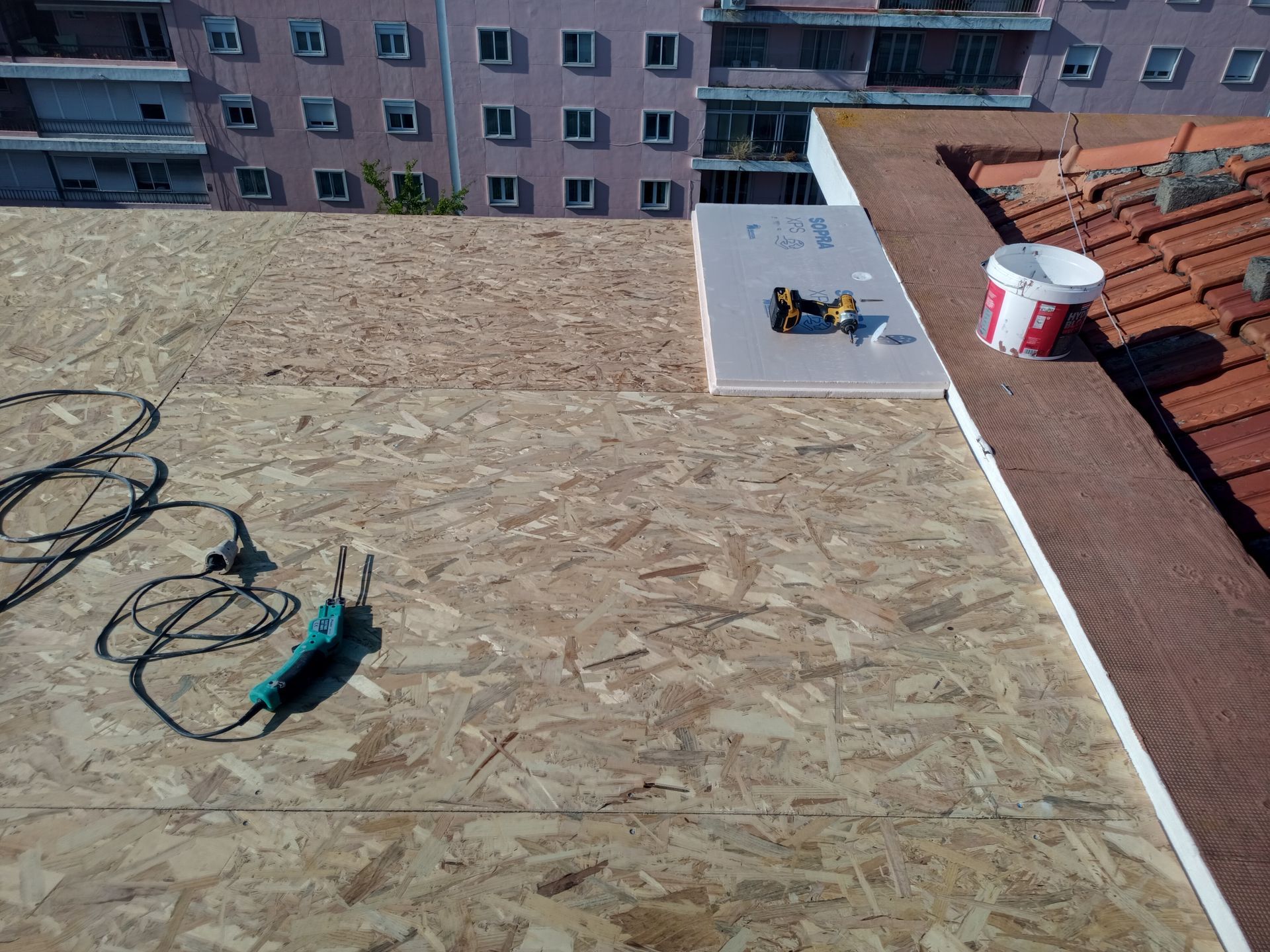 Construção no telhado: placas de OSB, isolamento, ferramentas e um balde de tinta estão próximos a um telhado de telhas vermelhas e a um prédio de vários andares.