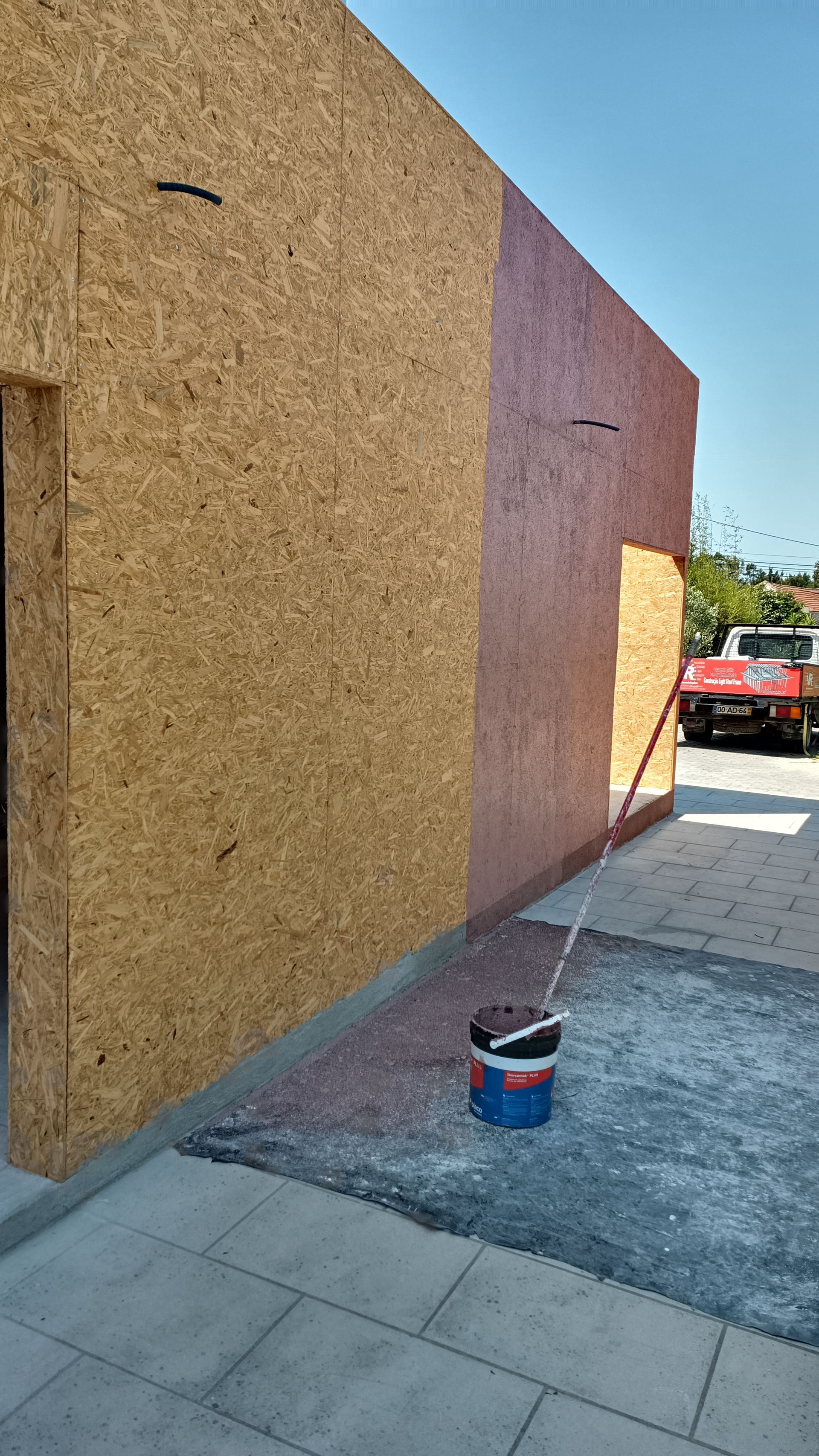 Fachada de um prédio com painéis de madeira, uma porta e um balde de tinta. Um carro vermelho está ao fundo.
