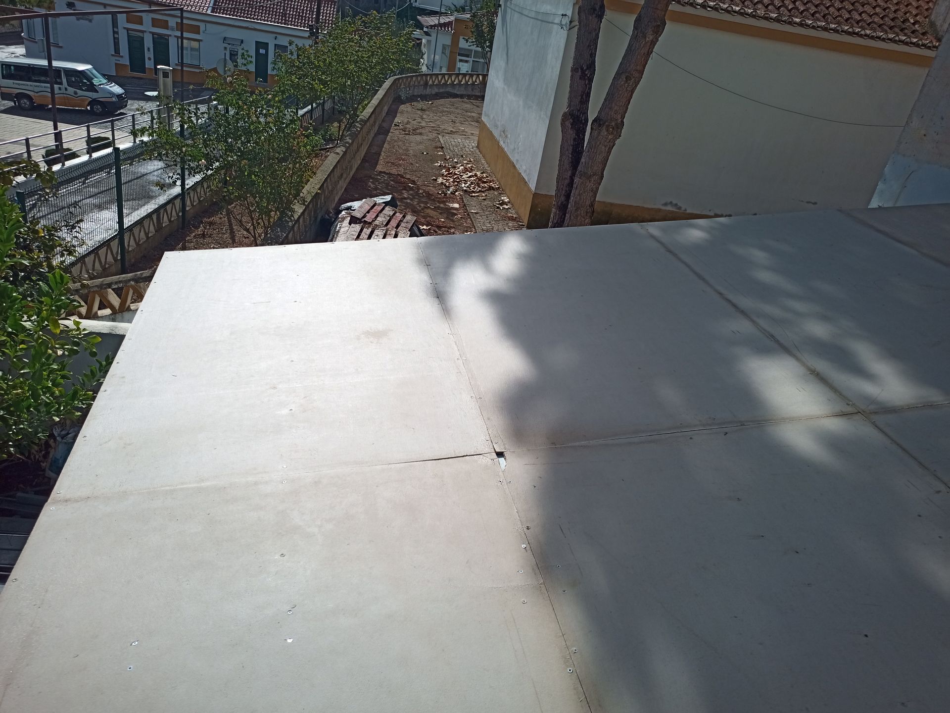 Vista de um telhado plano e claro com juntas visíveis, árvores e edifícios ao fundo.